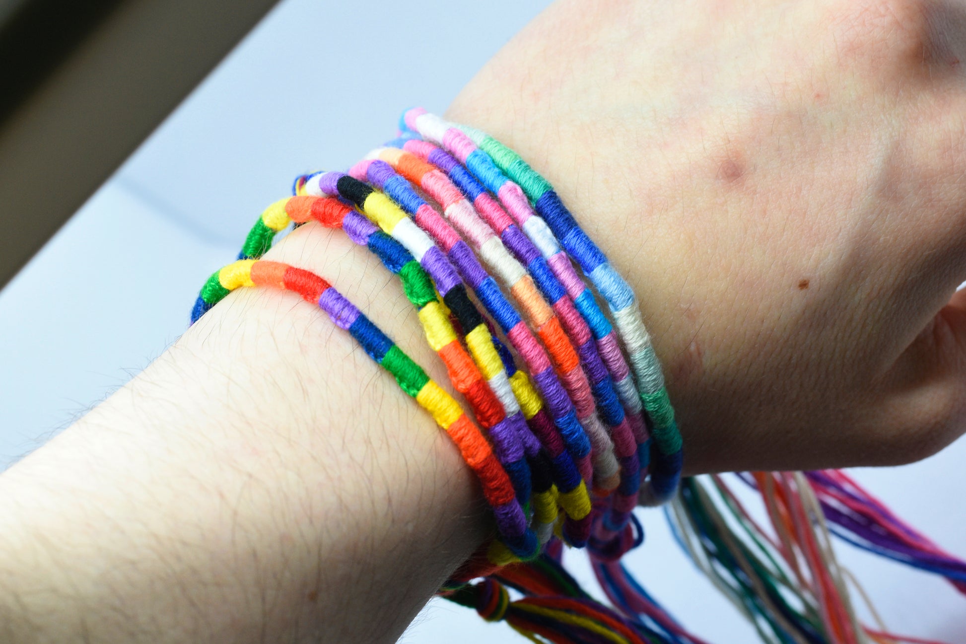 A collection of handmade pride bracelets made using embroidery thread in various pride flag colours. The flags shown include: rainbow, non-binary, bisexual, trans, lesbian and mlm. The bracelets are shown modelled on Alli's wrist.