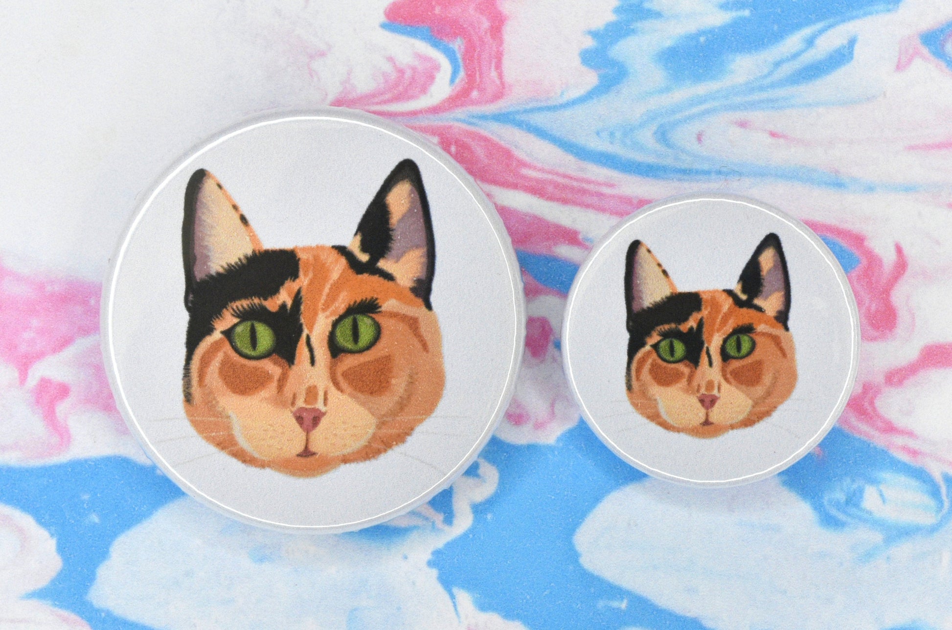 Two badges in sizes medium and small respectively, each featuring an off-white background with an illustration of the face of a ginger and black calico/tortoiseshell cat with green eyes.