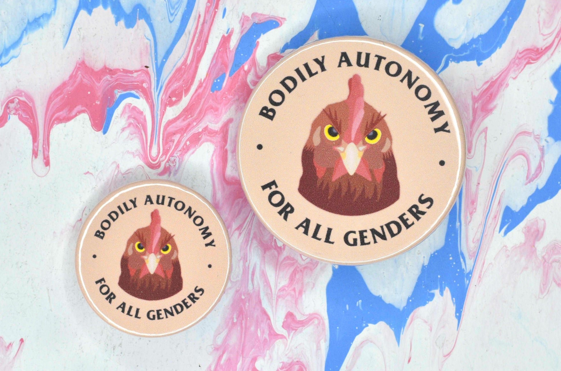 Two badges in sizes medium and small respectively, each featuring a peach coloured background with an illustration of a chicken with an angry expression. There is text surrounding the chicken reading "bodily autonomy for all genders".