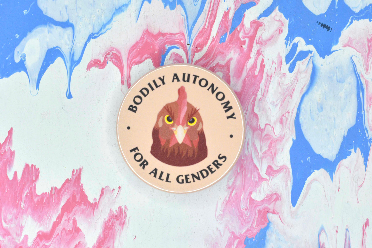 A badge in size medium featuring a peach coloured background with an illustration of a chicken with an angry expression. There is text surrounding the chicken reading "bodily autonomy for all genders".