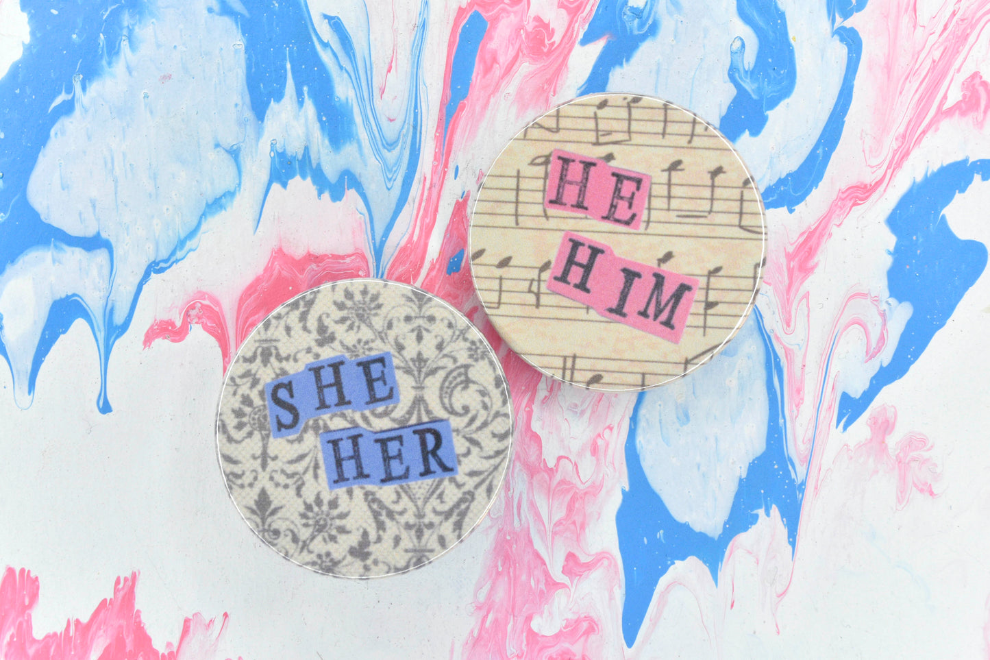 Two collage pronoun pins. On the left is a pin badge featuring an off-white background with a lace pattern in dark grey. Overlaid are hand cut pieces of purple paper with hand stamped uppercase black lettering reading "she" and "her". On the right is a pin badge featuring an off-white background with bars of music in black ink. Overlaid are hand cut pieces of pastel pink paper with hand stamped uppercase black lettering reading "he" and "him". 