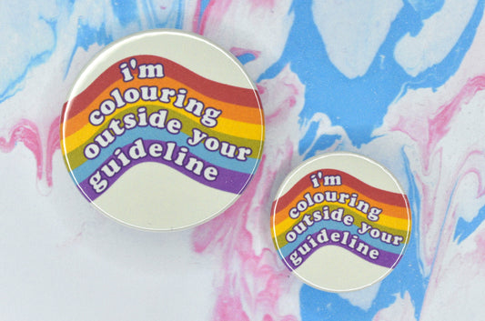 Two badges in sizes medium and small respectively, each featuring a pale yellow background with a curving rainbow design running across it. There is white bubble-like text overlaid reading "I'm colouring outside your guideline".