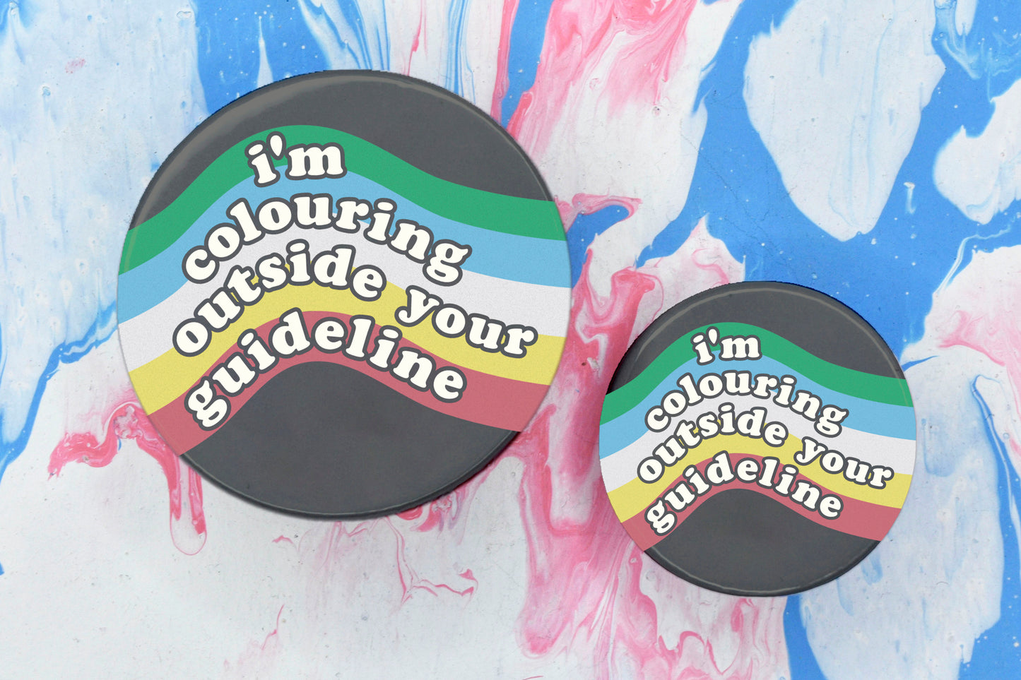 Two badges in sizes medium and small respectively, each featuring a dark grey background with a curving rainbow design in colours green, blue, white, yellow and red running across it. There is white bubble-like text overlaid reading "I'm colouring outside your guideline".