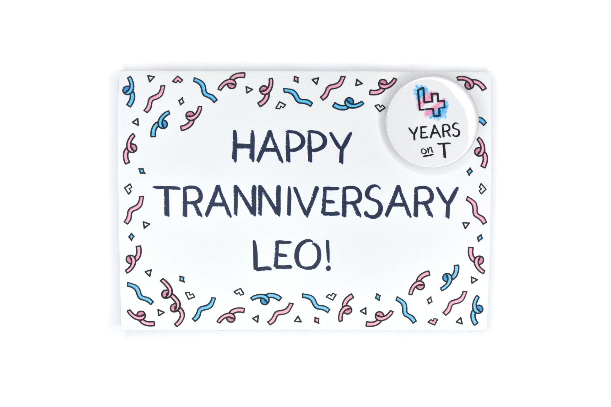 A photograph of a greetings card in landscape orientation featuring uppercase black text in a hand-drawn font which reads "Happy Tranniversary Leo!". There is illustrated confetti forming a border around the edges of the card in pink, blue and white. In the top right hand corner of the card there is a circular button badge with a white background and hand-drawn text reading "4 years on T" with the number being coloured in blue, pink and white.