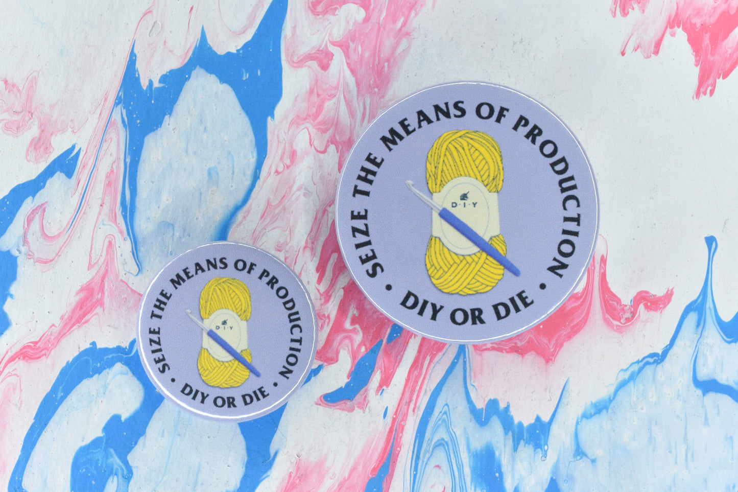 Two circular pin badges in sizes medium and small, each with a purple background and an illustration of a ball of yellow yarn with a purple handled crochet hook. There is uppercase black serif text surrounding this reading "seize the means of production - DIY or die".