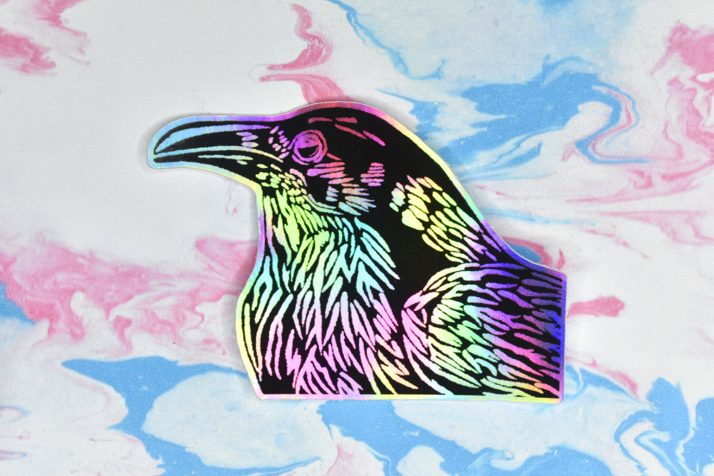 A sticker featuring a crow illustration in black ink on an iridescent holographic background. The holographic effect produces a metallic rainbow of colour across the surface of the sticker.