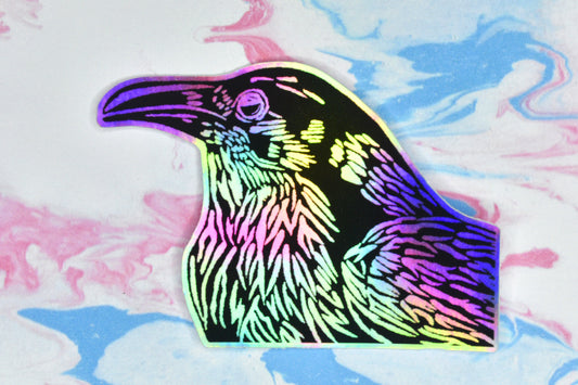 A sticker featuring a crow illustration in black ink on an iridescent holographic background. The holographic effect produces a metallic rainbow of colour across the surface of the sticker.