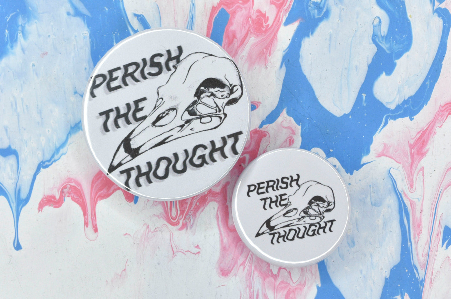 Two pin badges in sizes medium and small, both featuring a white background with a black and white illustration of a bird skull with text surrounding it that reads "perish the thought". 