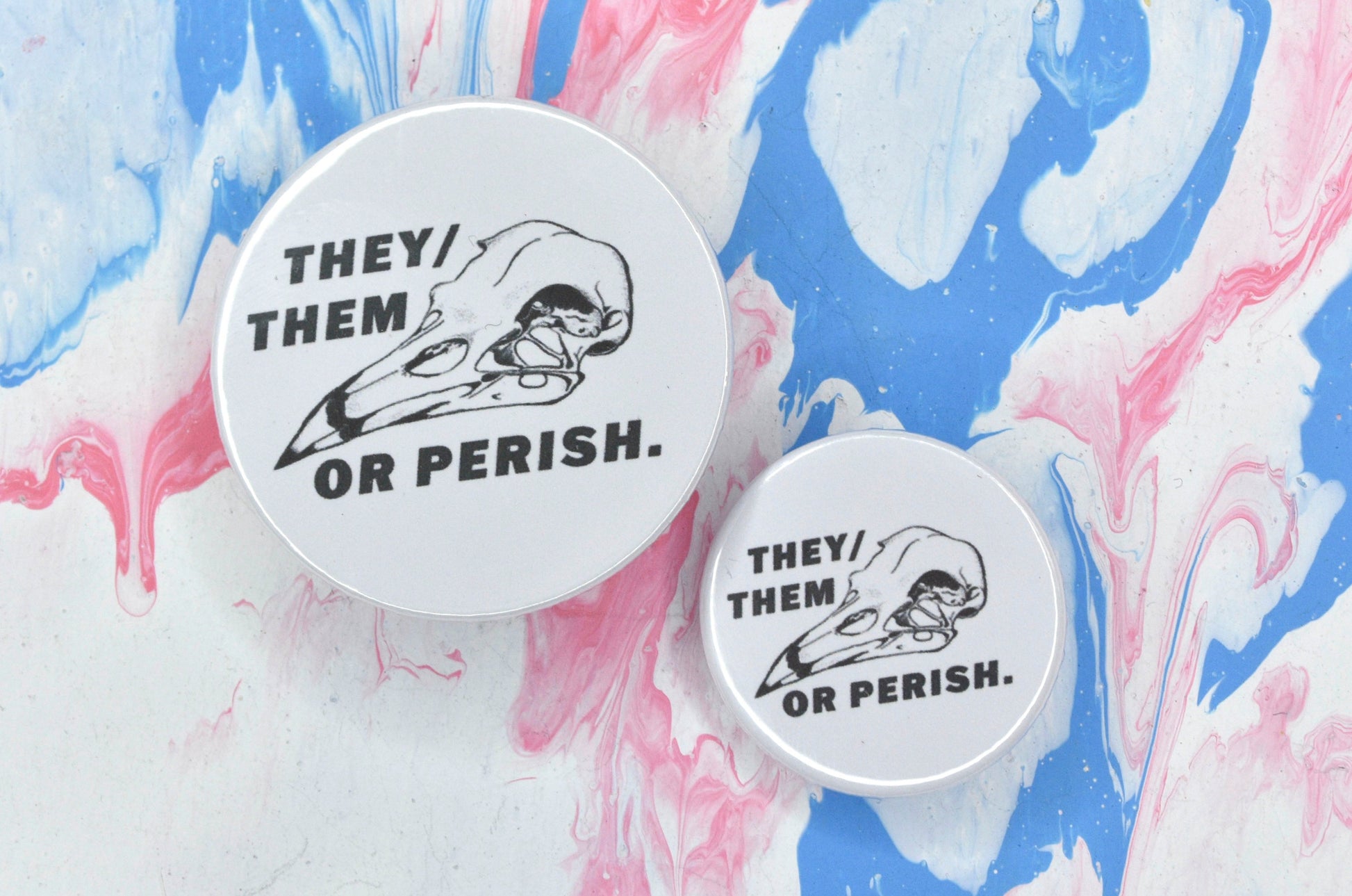 Two pin badges in sizes medium and small, both with a white background featuring a black and white illustration of a bird skull and text reading "they/them or perish".