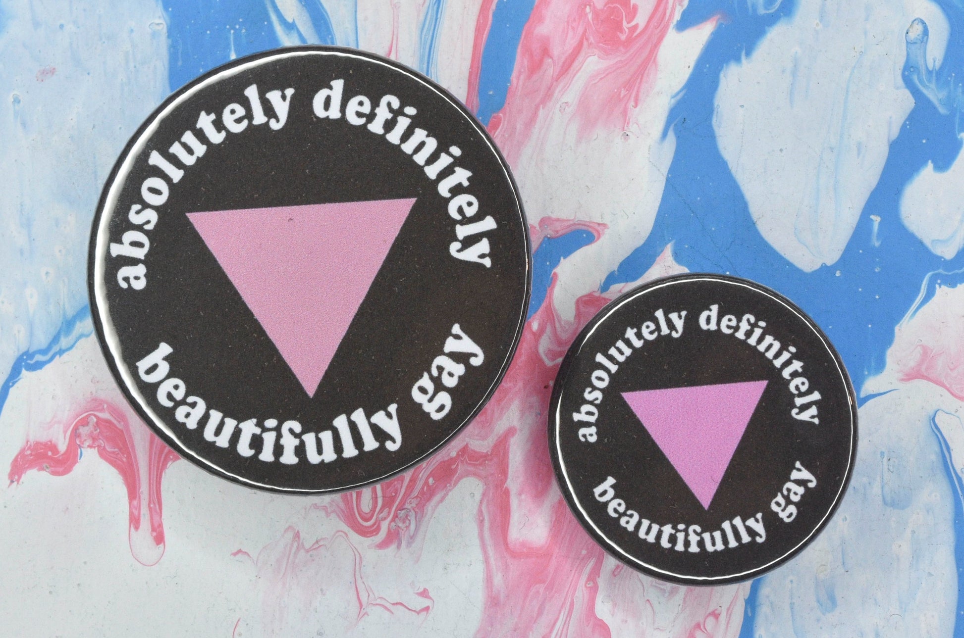 Two pin badges in sizes medium and small, both featuring a black background with a pink triangle in the centre and white text around the outside reading "absolutely definitely beautifully gay".