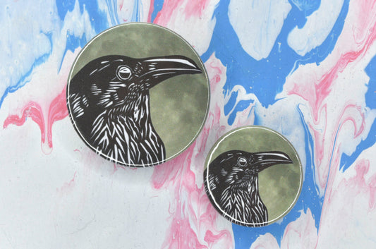 Two badges in sizes medium and small respectively, each featuring a black and white illustration showing the side profile of a crow from the breast up. The background is a yellowish grey close-up image of the moon.