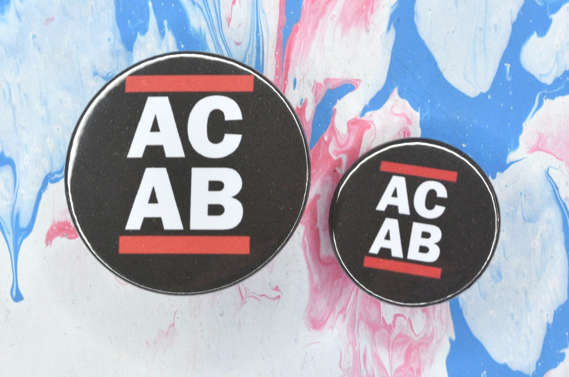 A circular pin badge design in sizes small and medium, both with a black background, two rows of bold white upper case text and a solid red line above and below the text. The text on the pin reads "ACAB", with the letters split between the two rows of text.