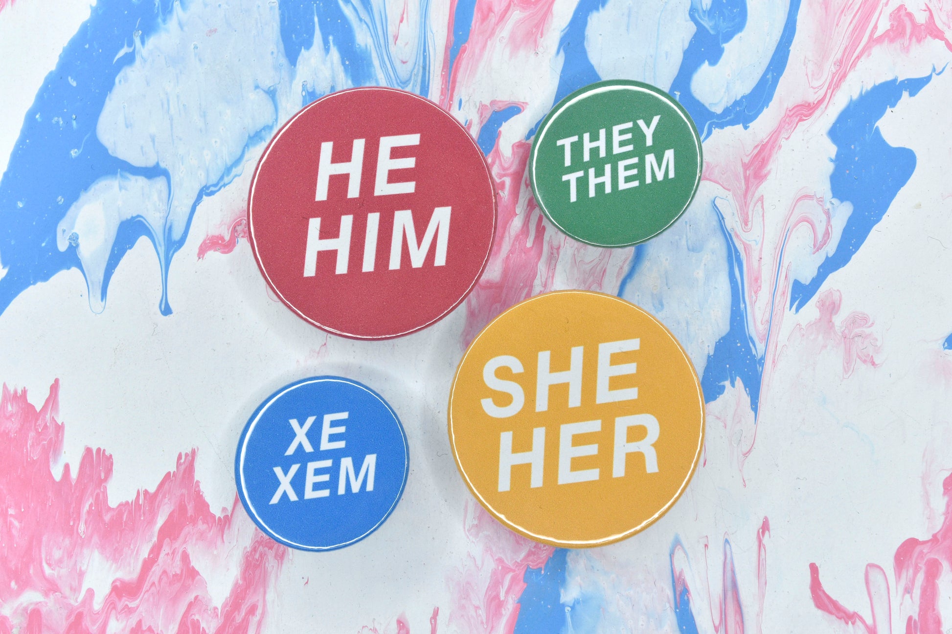 Four pronoun pins, each with a solid colour background and white text featuring various sets of pronouns. The pins are shown in dark pink with pronouns "he/him", yellow with pronouns "she/her", blue with "xe/xem" and green with "they/them".