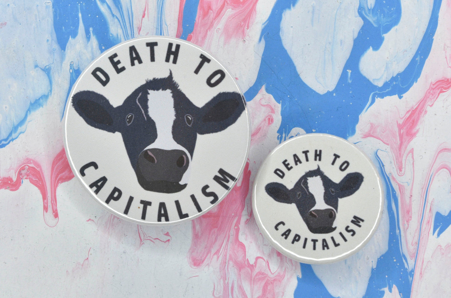 Two pin badges in sizes medium and small featuring an illustration of a cow with text reading "death to capitalism" on a white background.