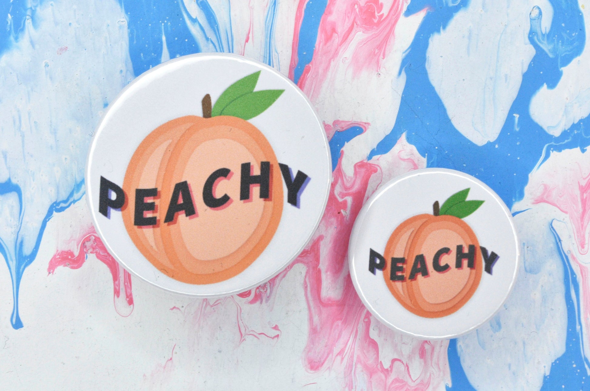 Two pin badges in sizes medium and small, both featuring a white background with an illustration of a peach in the centre. There is text across the centre of the peach reading "peachy".