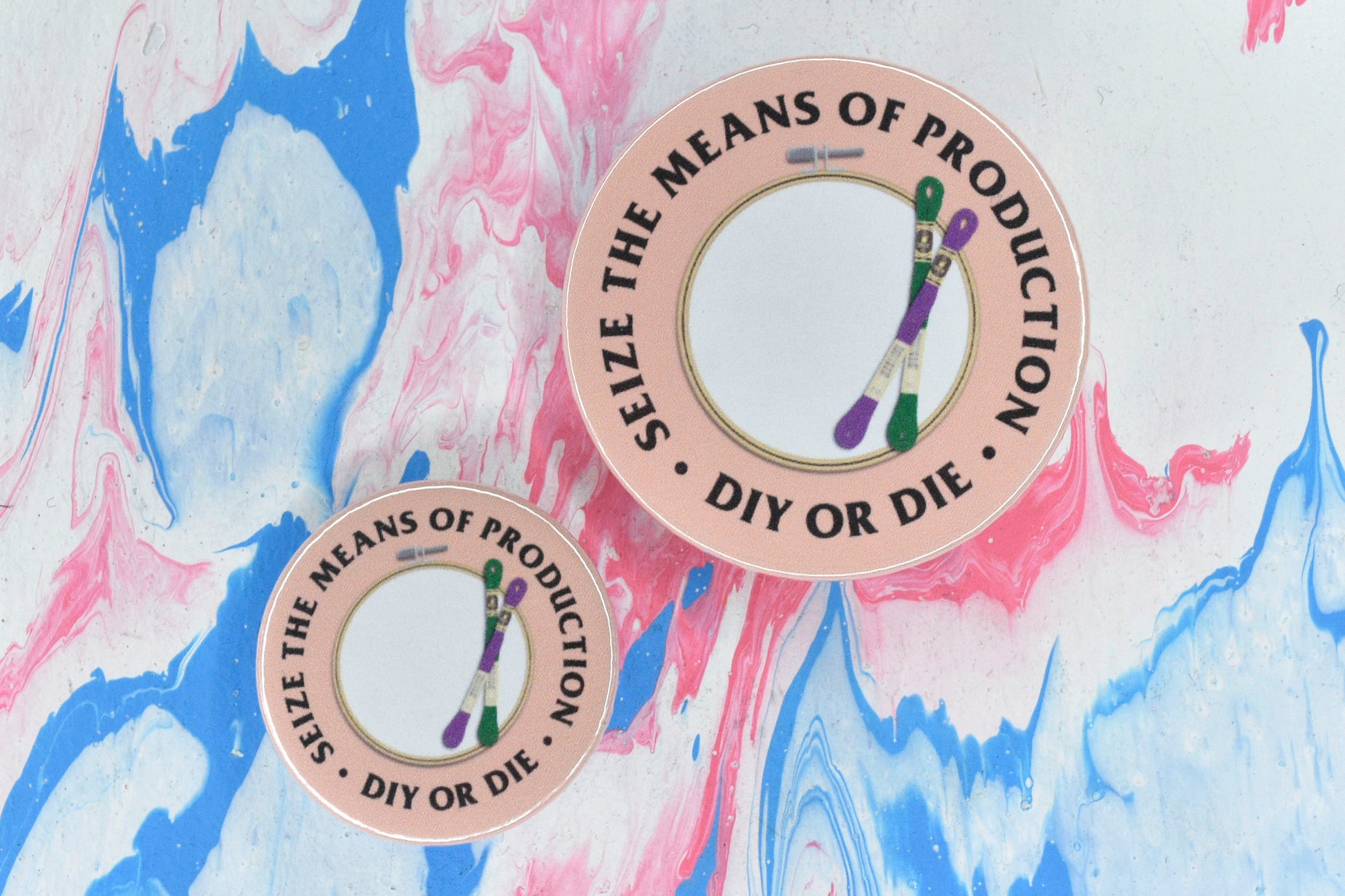 Two circular pin badges in sizes medium and small, each with a peach background and an illustration of an embroidery hoop with two skeins of green and purple thread. There is uppercase black serif text surrounding this reading "seize the means of production - DIY or die".