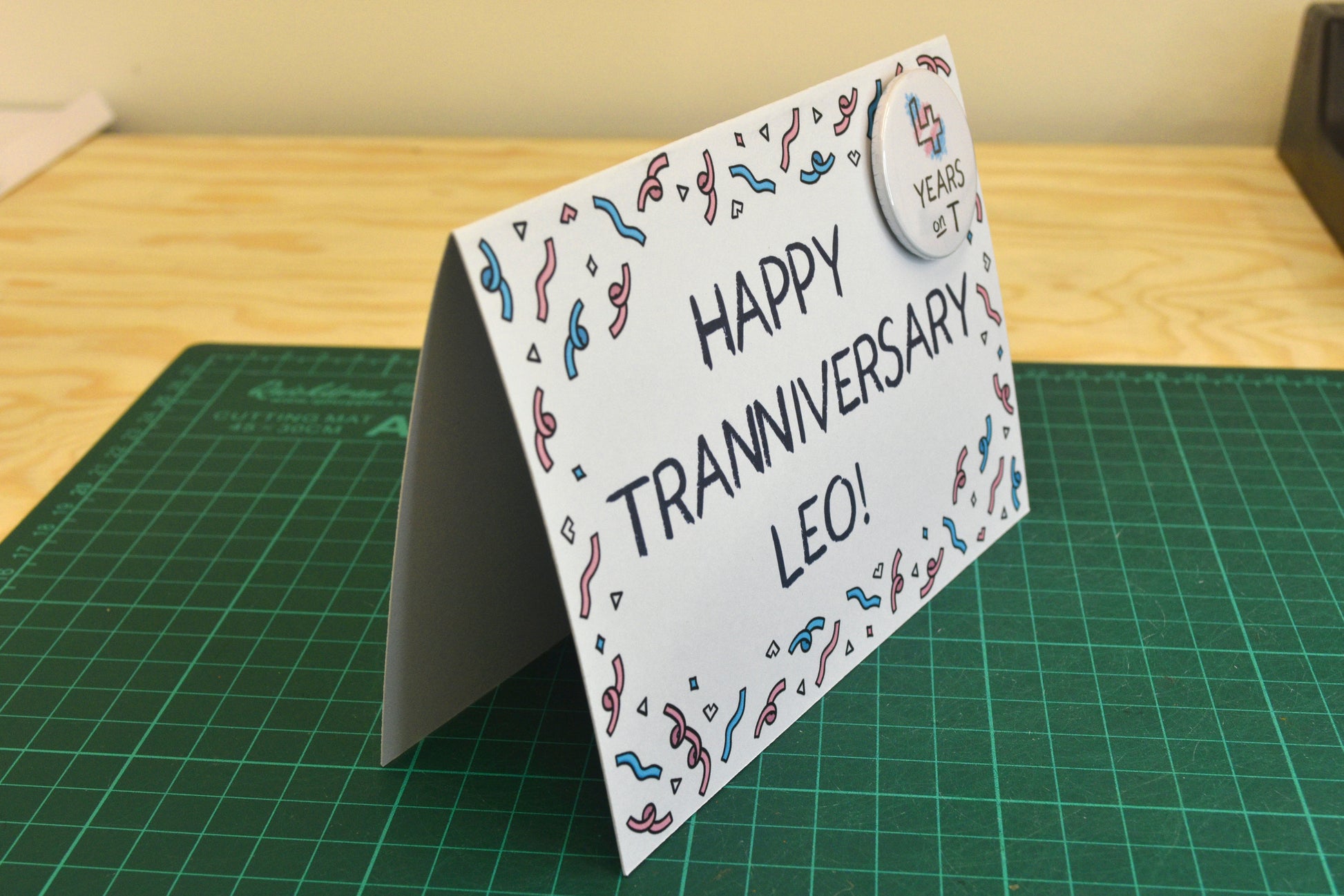 A photograph of a greetings card in landscape orientation featuring uppercase black text in a hand-drawn font which reads "Happy Tranniversary Leo!". There is illustrated confetti forming a border around the edges of the card in pink, blue and white. In the top right hand corner of the card there is a circular button badge with a white background and hand-drawn text reading "4 years on T" with the number being coloured in blue, pink and white. The card is standing up in a tent shape.