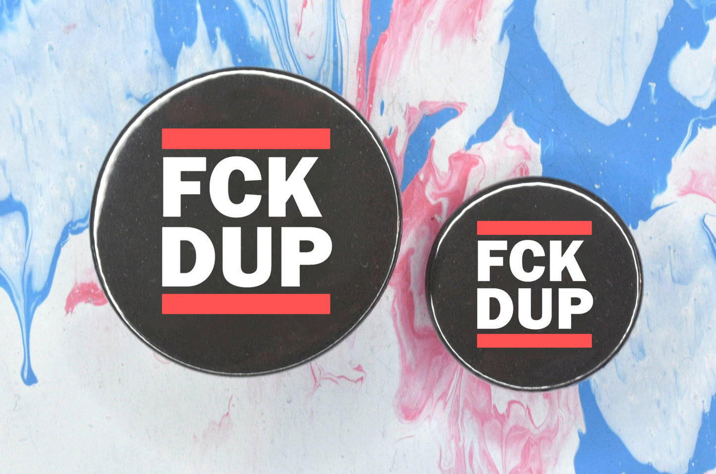 Two circular pin badges in sizes medium and small each with a black background, two rows of bold white upper case text and a solid red line above and below the text. The top row reads "fck" and the bottom row reads "dup", i.e. "fuck the DUP".