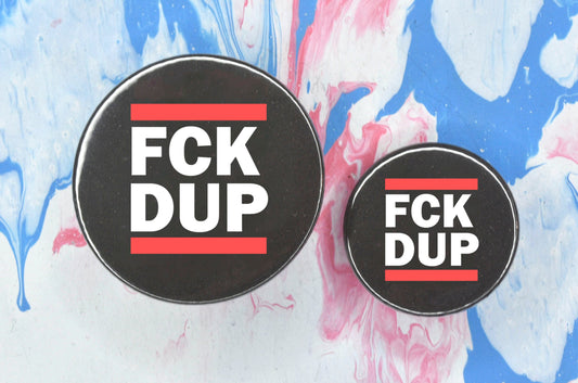 Two circular pin badges in sizes medium and small each with a black background, two rows of bold white upper case text and a solid red line above and below the text. The top row reads "fck" and the bottom row reads "dup", i.e. "fuck the DUP".