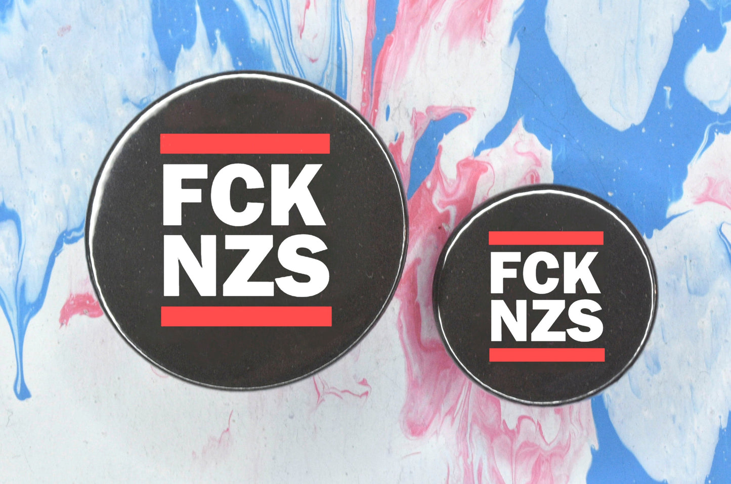 Two circular pin badges in sizes medium and small each with a black background, two rows of bold white upper case text and a solid red line above and below the text. The top row reads "fck" and the bottom row reads "nzs".