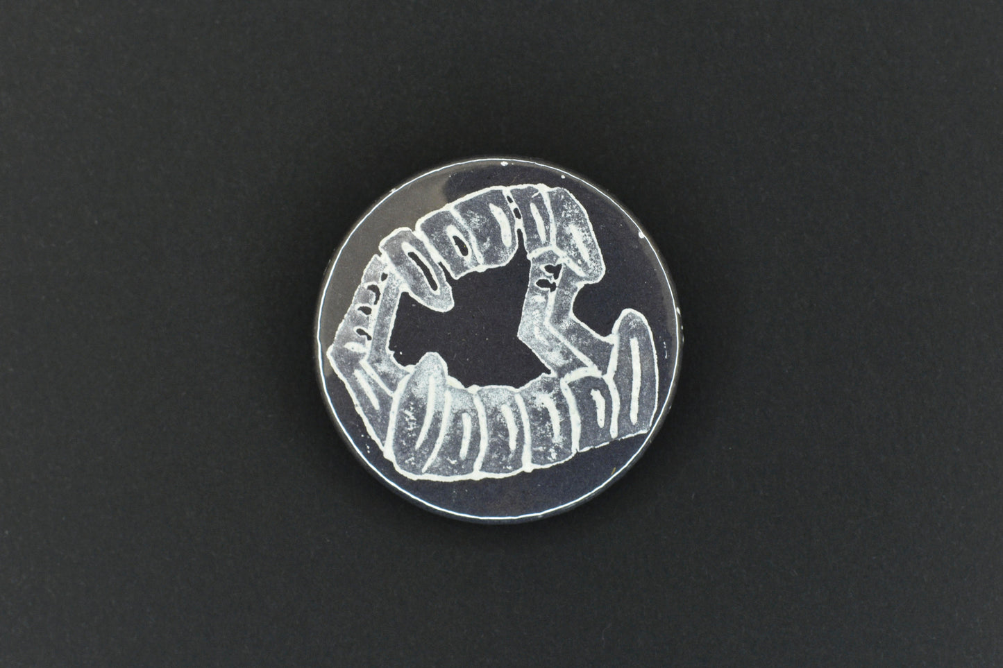 A single button badge featuring a black background with an original lino print stamped fang design in white ink.