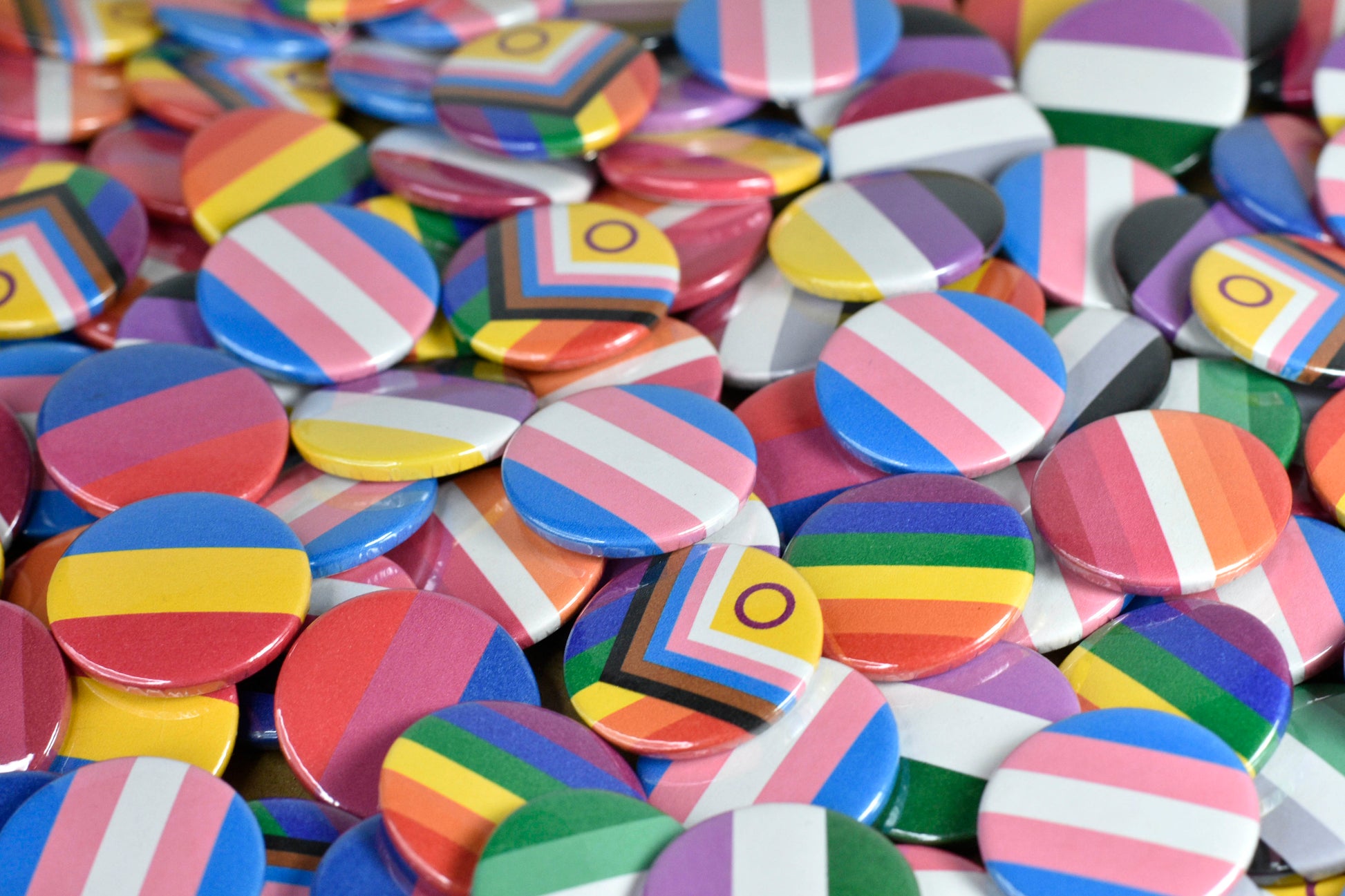 A large pile of small circular pin badges featuring a range of pride flags including progress pride, rainbow, trans, non-binary, asexual, bisexual, pansexual, lesbian and aromantic.