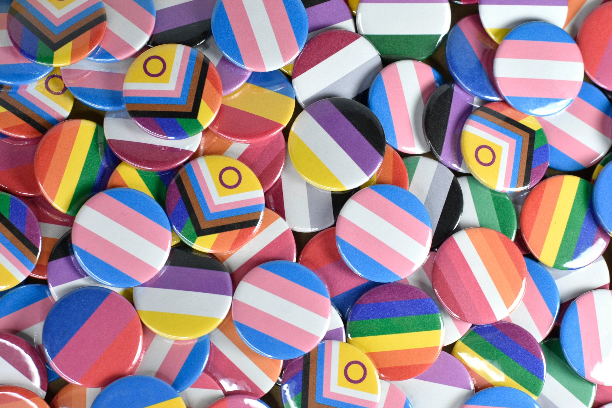 A large pile of small circular pin badges featuring a range of pride flags including progress pride, rainbow, trans, non-binary, asexual, bisexual, pansexual, lesbian and aromantic.