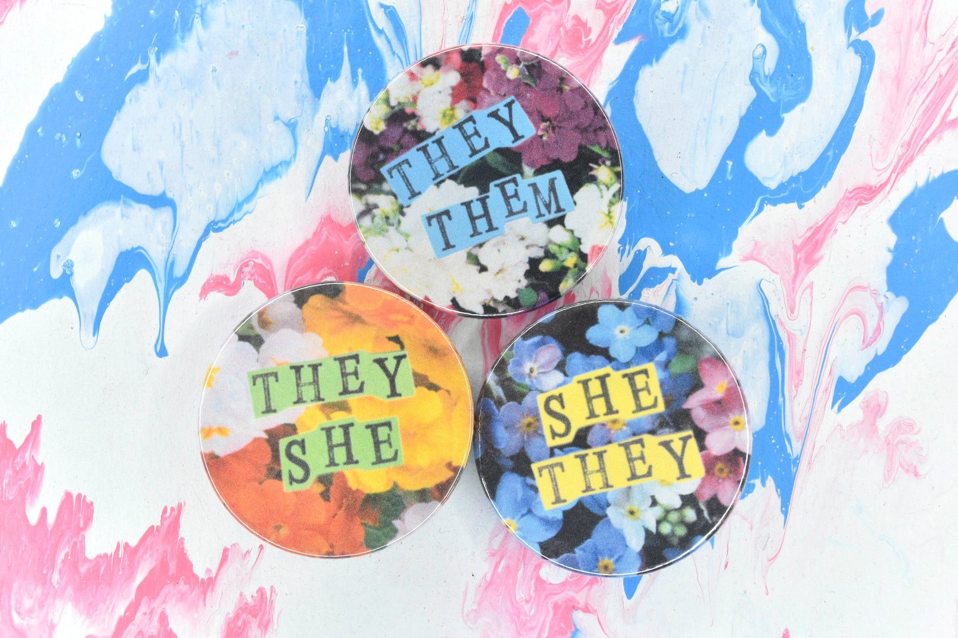 A collection of three pronoun badges featuring different floral backgrounds and pronouns in hand-stamped lettering. The options shown are a pink and white flowers with blue pastel paper reading "they/them"; orange, yellow and white flowers with green paper reading "they/she"; and blue, white and pink flowers with yellow paper reading "she/they".