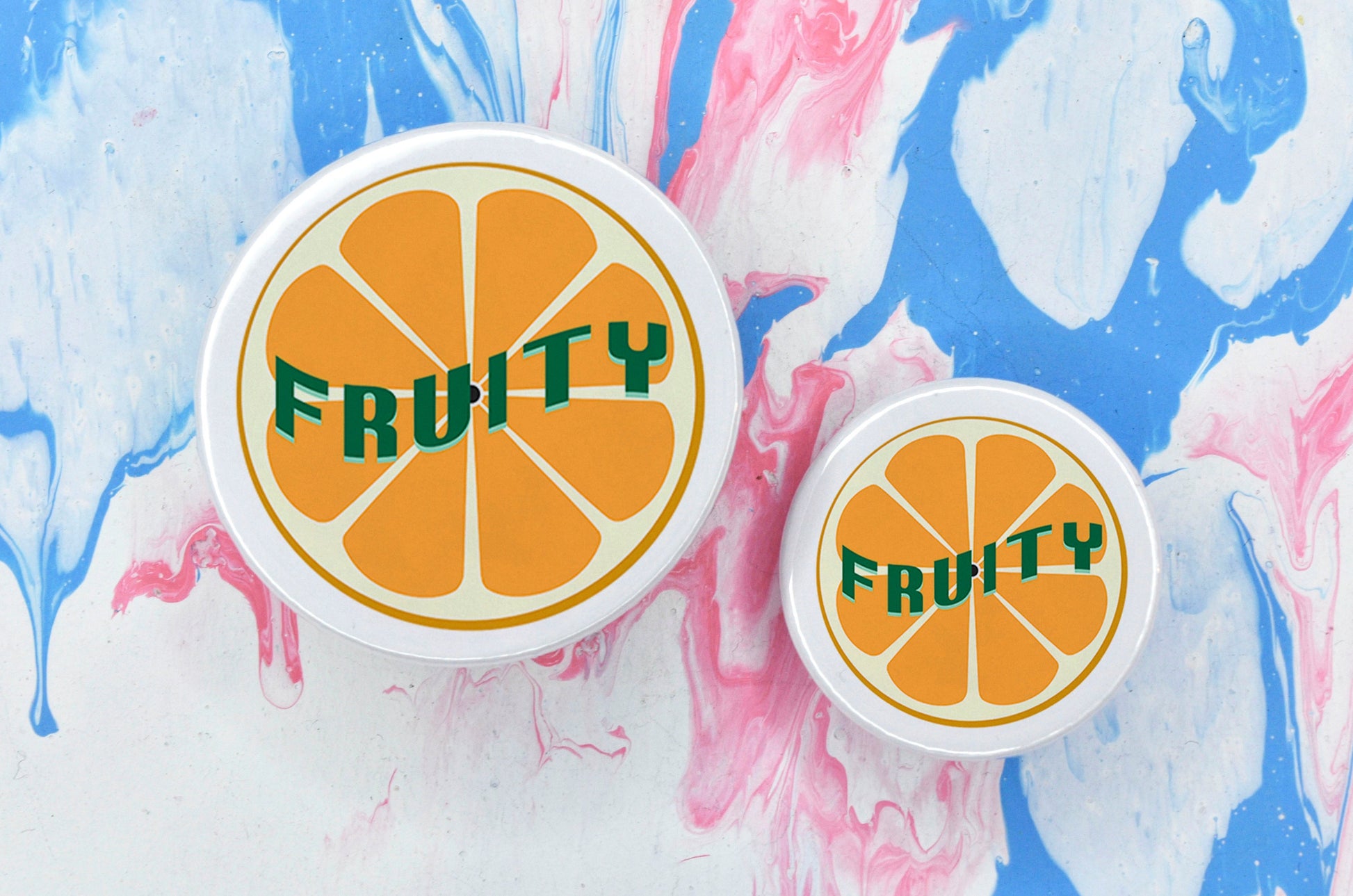 Two badges in sizes medium and small respectively, each featuring a white background with an illustration of an orange in the centre. There is uppercase green text across the centre of the orange reading "fruity".