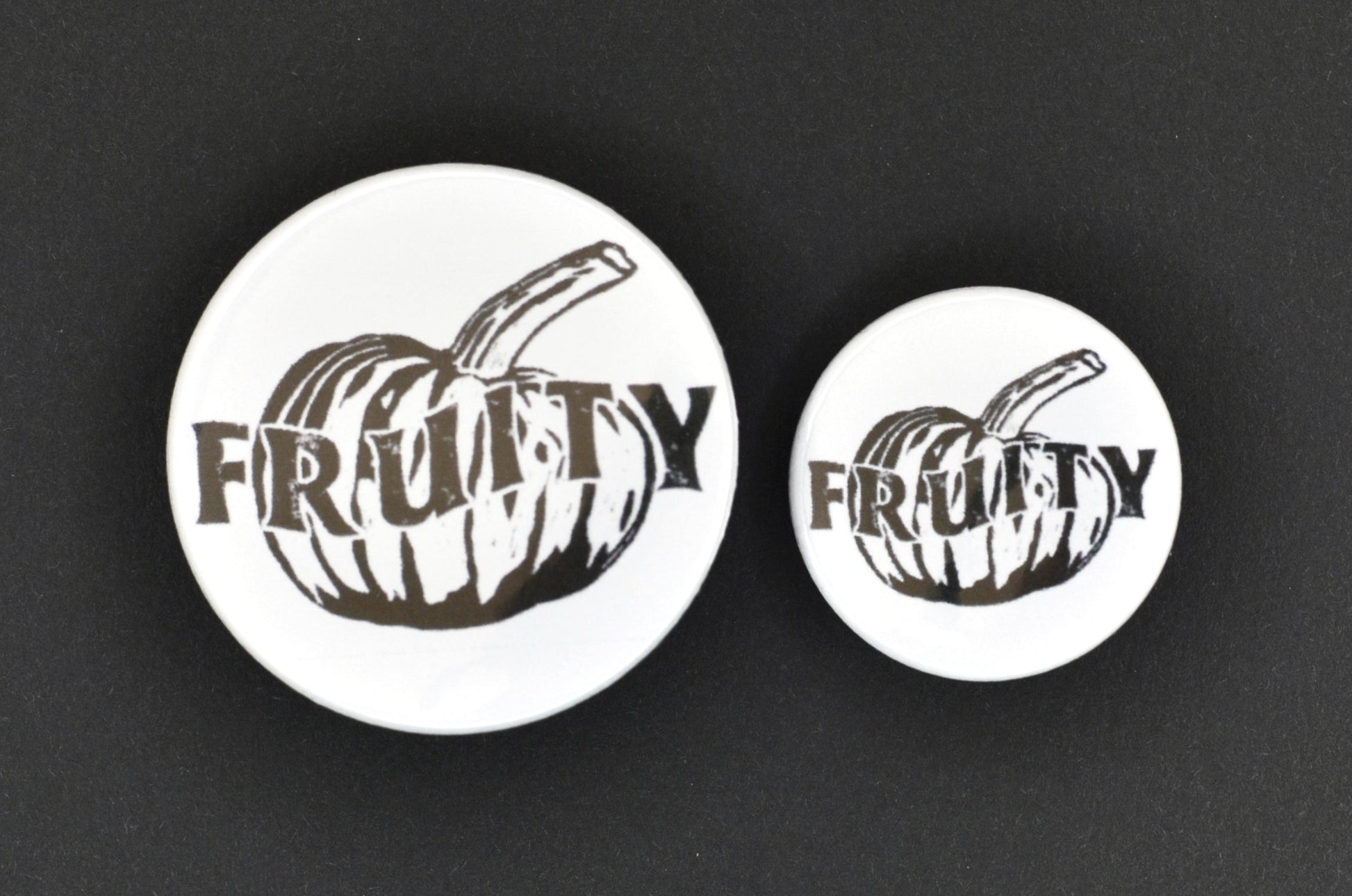 Two badges in sizes medium and small respectively, each featuring a white background with a black and white pumpkin in a lino print style in the centre. There is uppercase black text across the centre of the pumpkin reading "fruity".