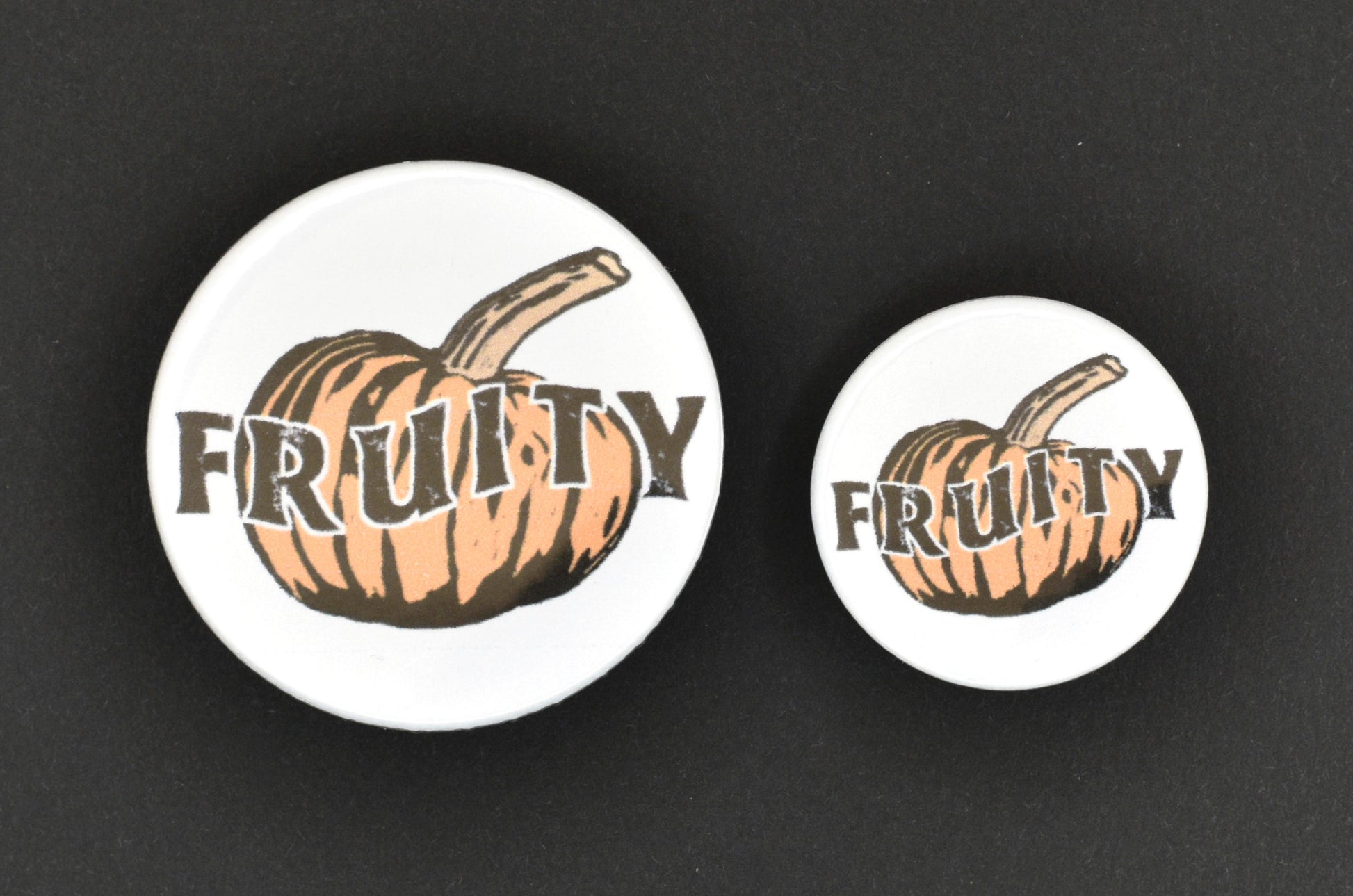 Two badges in sizes medium and small respectively, each featuring a white background with an orange pumpkin in a lino print style in the centre. There is uppercase black text across the centre of the pumpkin reading "fruity".