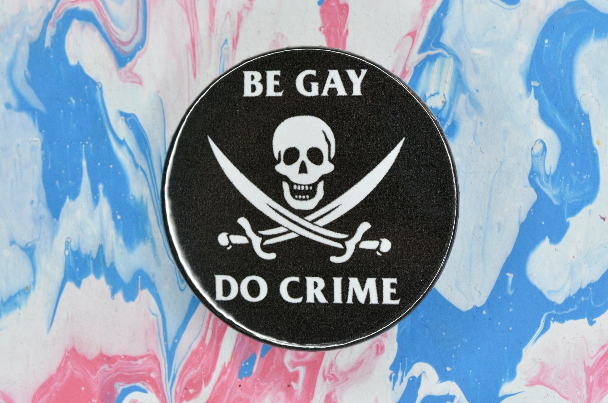 A black pin badge with white text reading "be gay" and the top and "do crime" at the bottom. In the centre is a skull and two swords (classic Jolly Rodger pirate flag)