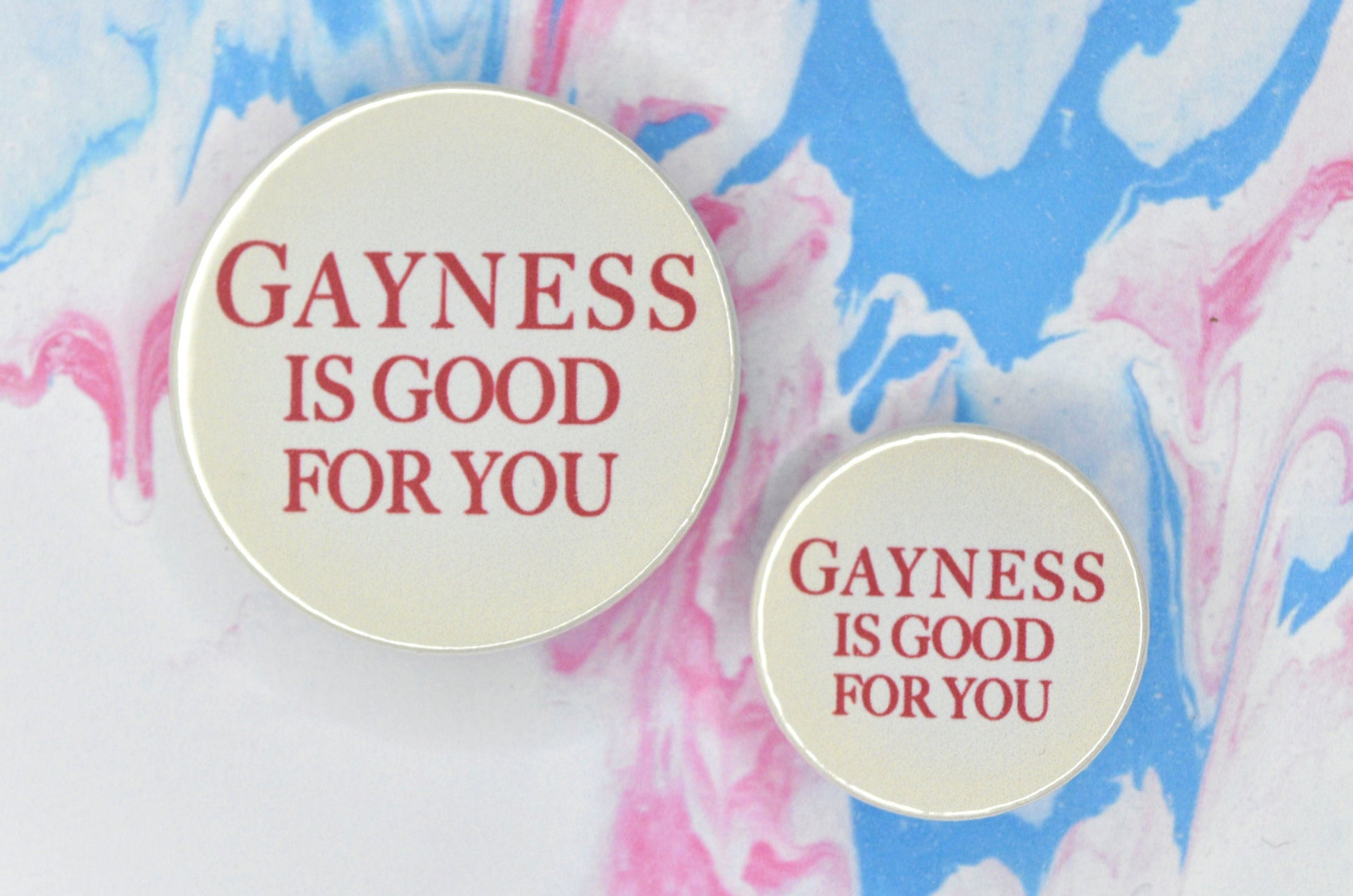 Badges in sizes small and medium featuring red serif typeface reading "gayness is good for you" on a light beige background.