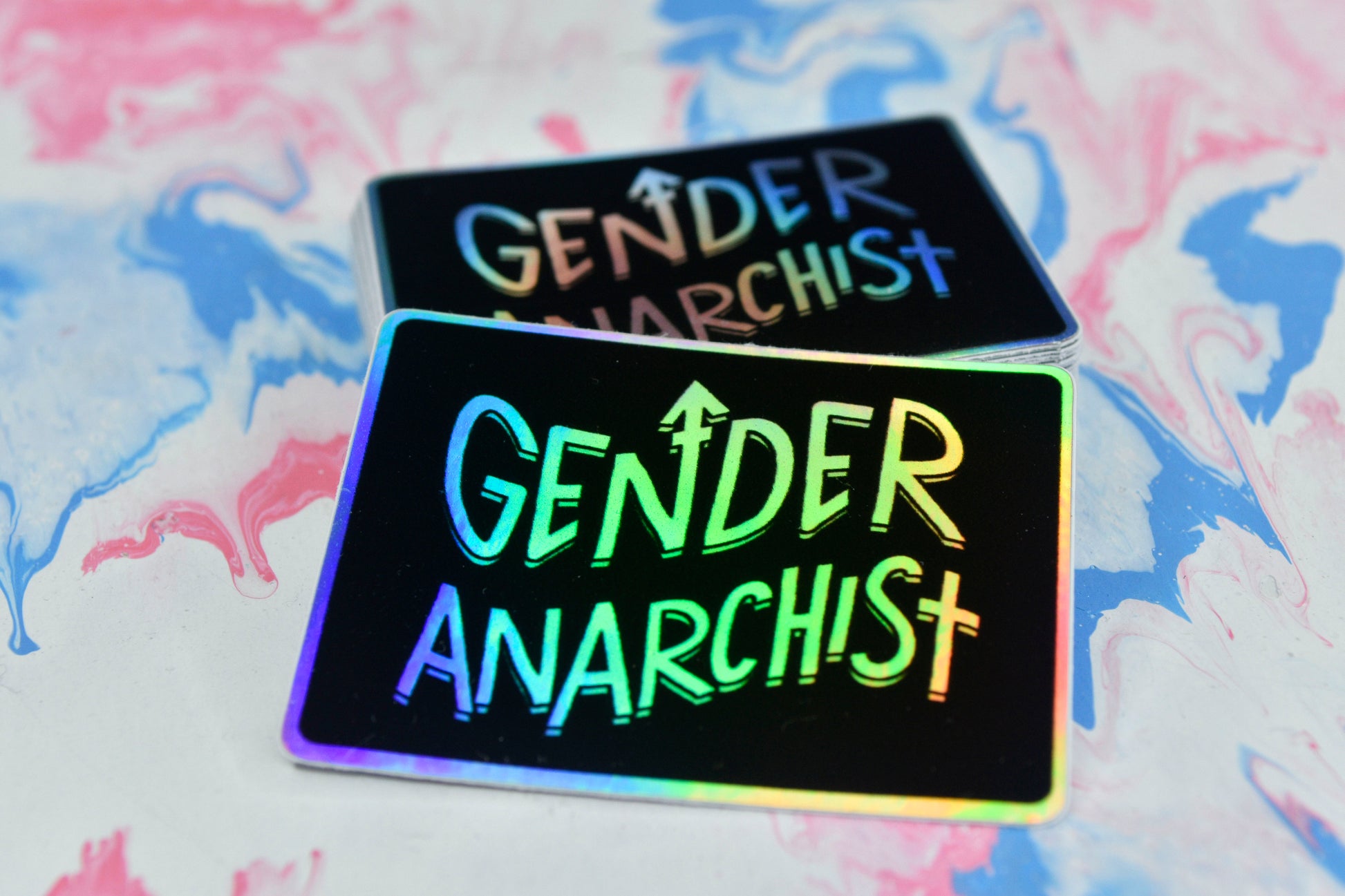 A rectangular holographic sticker with a black background and white holographic text reading "gender anarchist". It is shown propped against a small pile of the same stickers in the background.