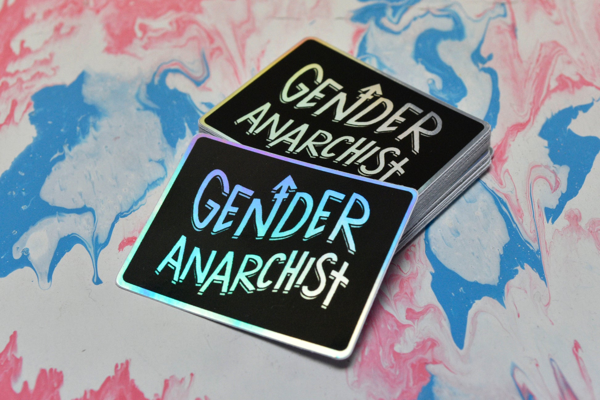 A rectangular holographic sticker with a black background and white holographic text reading "gender anarchist". It is shown propped against a small pile of the same stickers.