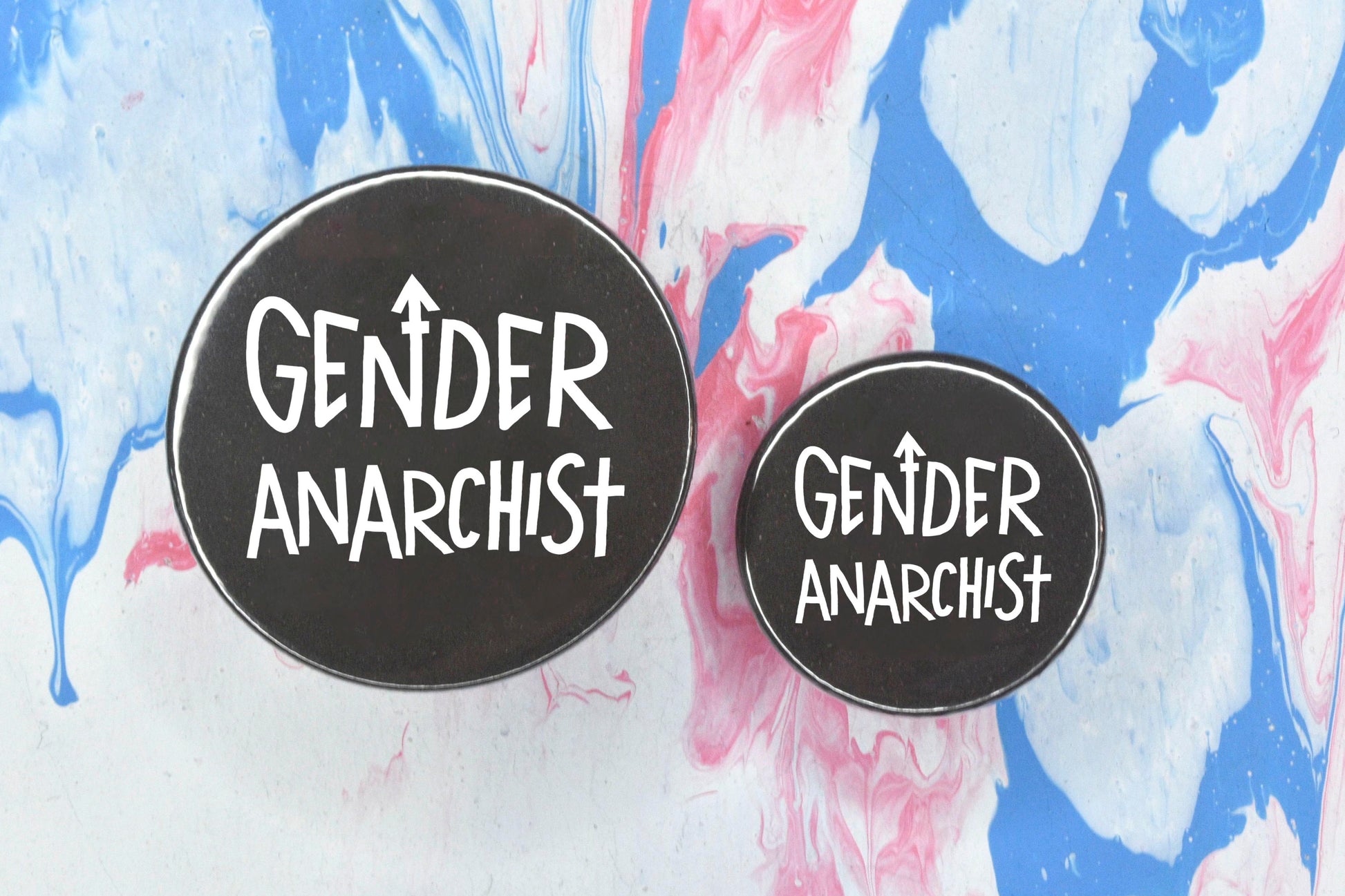 Two circular pin badges in sizes medium and small each with a black background and uppercase hand-drawn text in white which reads "gender anarchist".