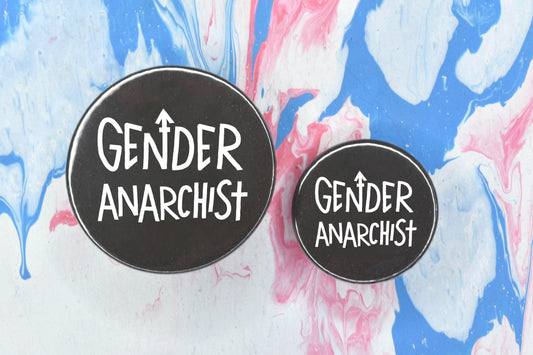 Two circular pin badges in sizes medium and small each with a black background and uppercase hand-drawn text in white which reads "gender anarchist".