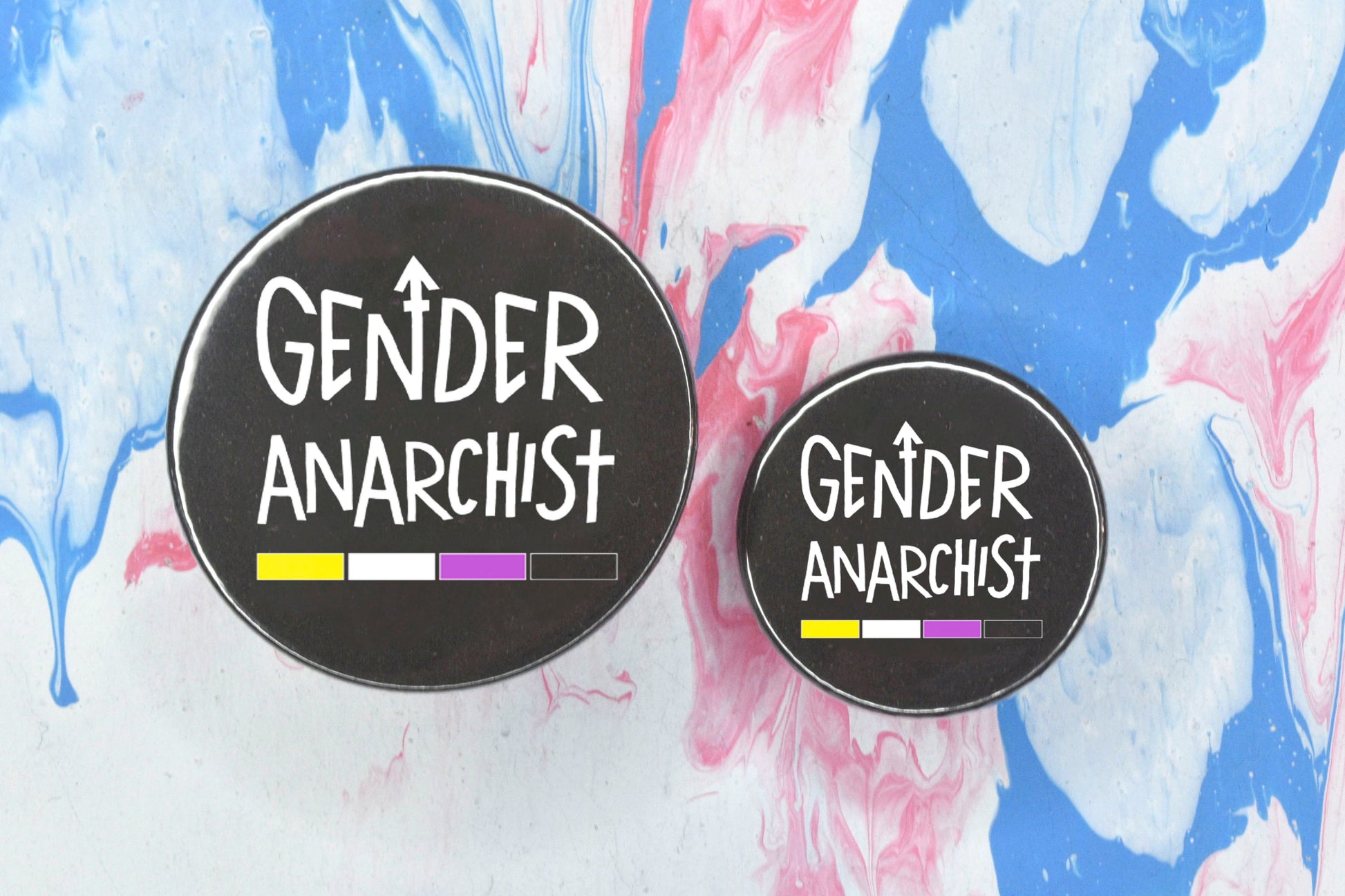 Two circular pin badges in sizes medium and small each with a black background and uppercase hand-drawn text in white which reads "gender anarchist". There is a band of colour along the bottom of the text which makes up the non-binary pride flag.