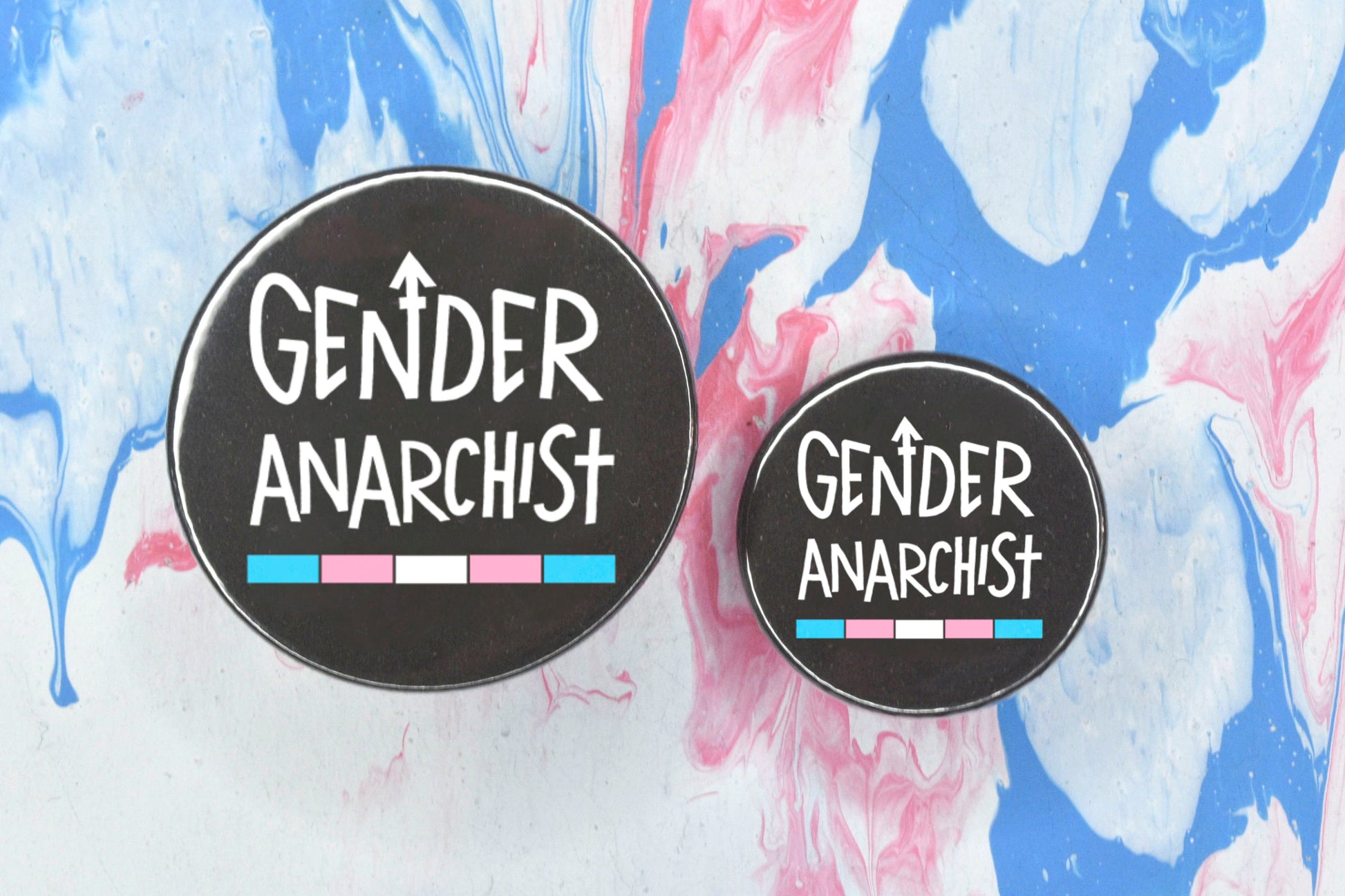 Two circular pin badges in sizes medium and small each with a black background and uppercase hand-drawn text in white which reads "gender anarchist". There is a band of colour along the bottom of the text which makes up the trans pride flag.