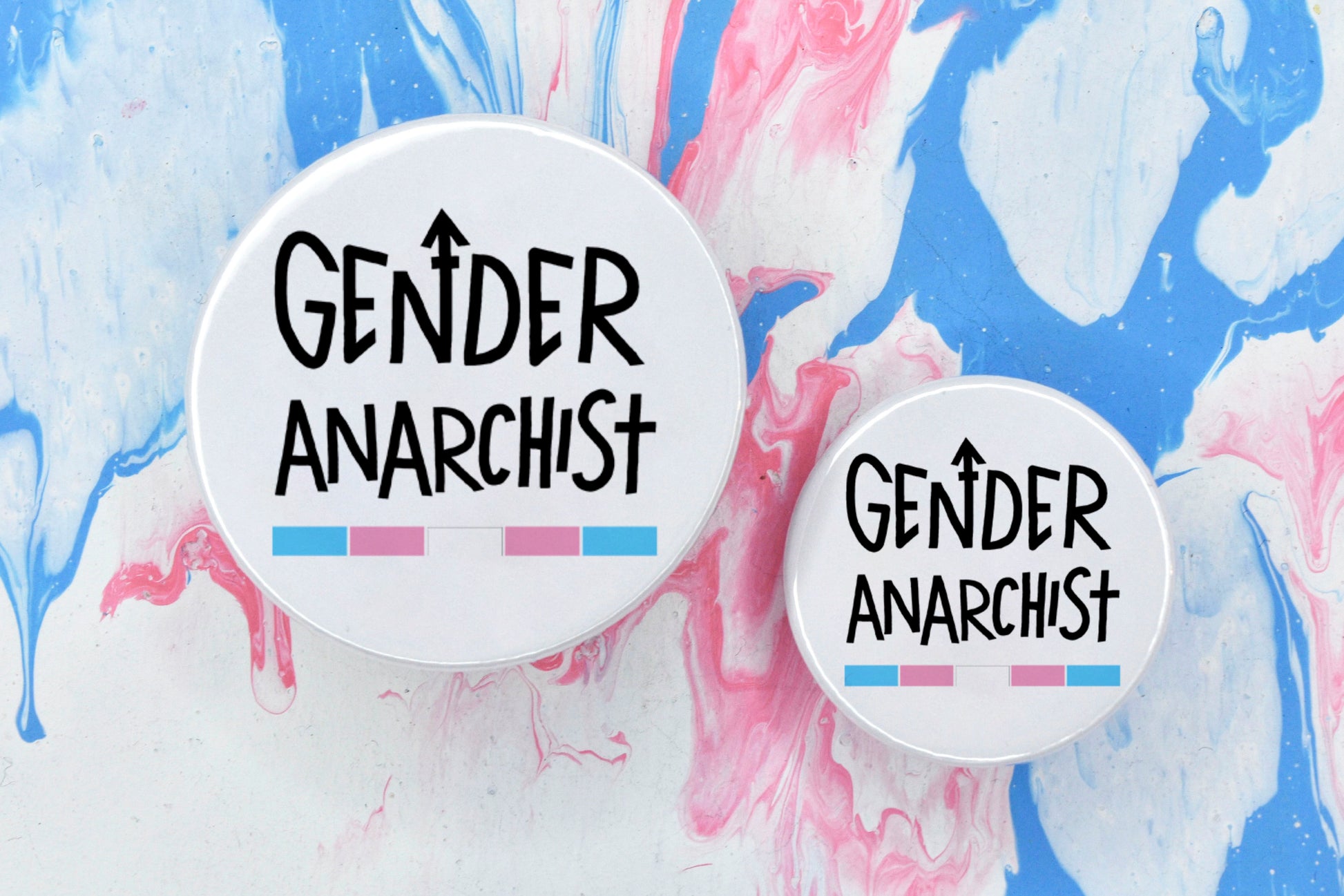 Two circular pin badges in sizes medium and small each with a white background and uppercase hand-drawn text in black which reads "gender anarchist". There is a band of colour along the bottom of the text which makes up the trans pride flag.