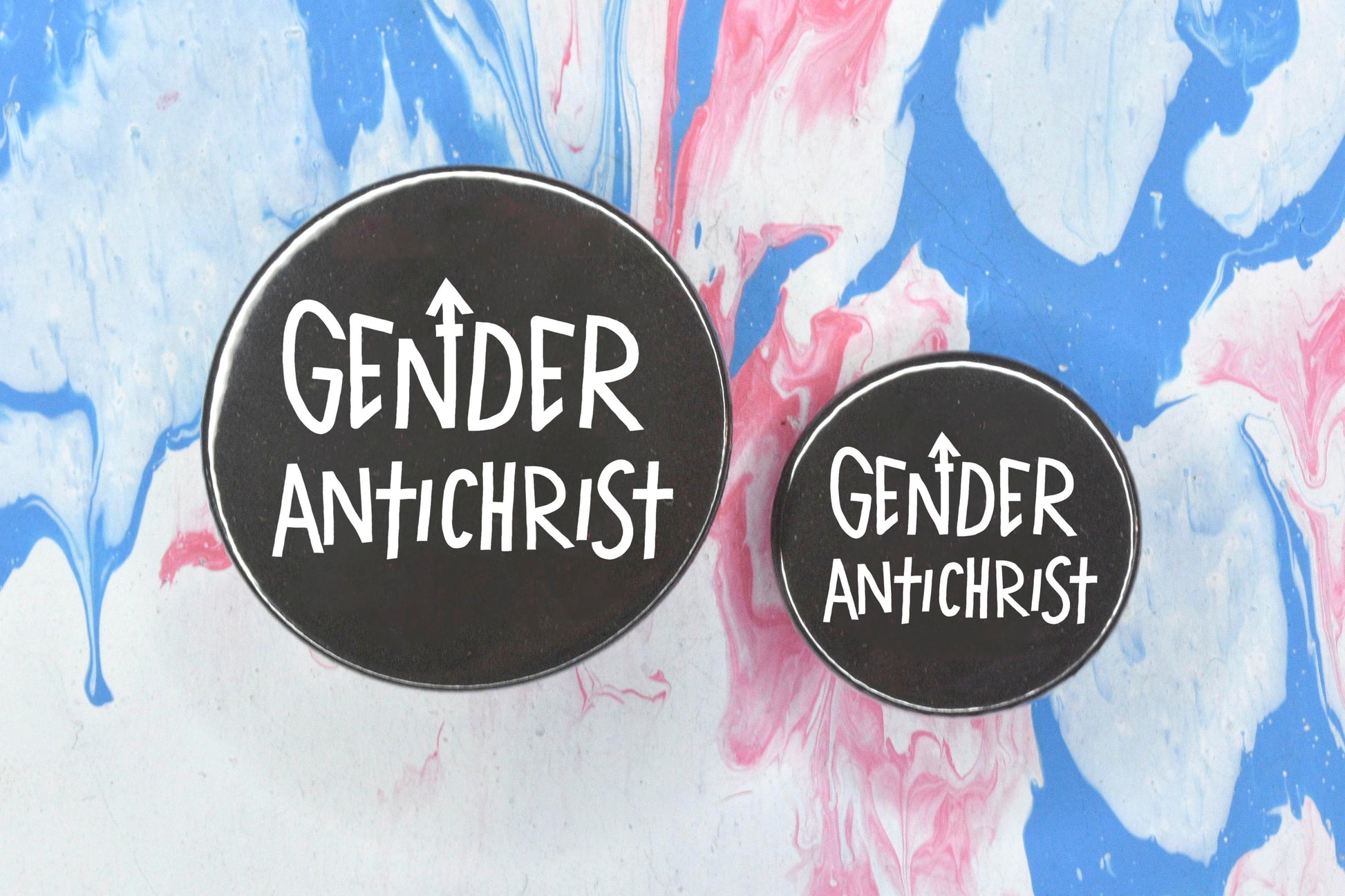 Two circular pin badges in sizes medium and small each with a black background and uppercase hand-drawn text in white which reads "gender antichrist".