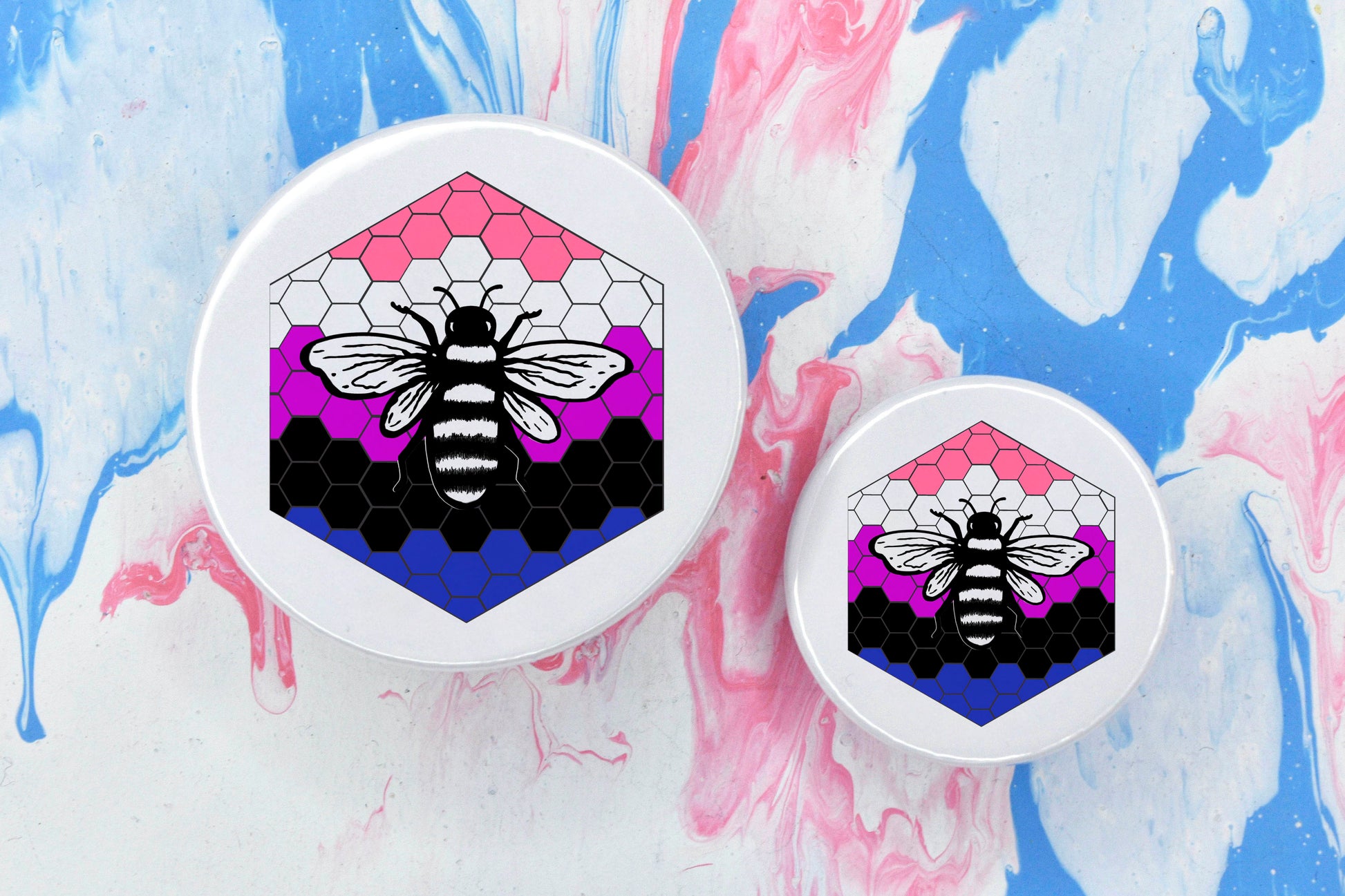 Two badges in sizes medium and small respectively, each featuring a white background with a black and white illustration of a bee overlaid on a bee hive pattern which is coloured in with the genderfluid pride flag colours (light pink, white, dark pink, black and blue).