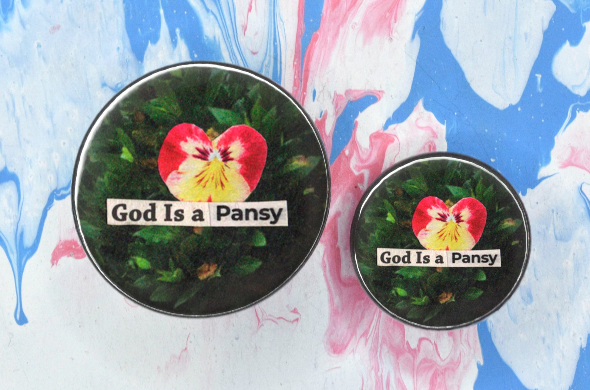 Two pin badges in sizes medium and small, both featuring a collage consisting of a small photo of a pink and yellow pansy flower on a green leafy background with newspaper print overlaid reading "god is a pansy".