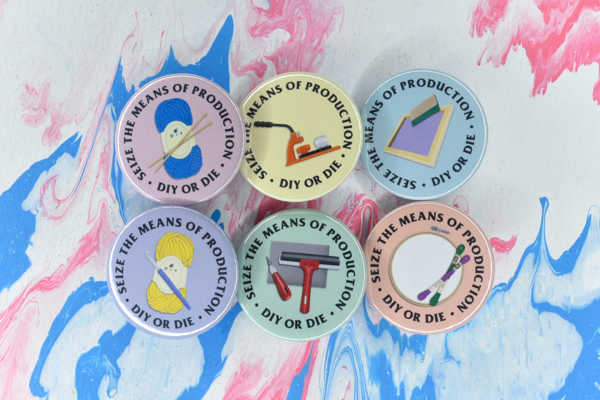 A group of six badges, each with a different coloured background, an illustration representing a different craft, and text reading "seize the means of production - DIY or die". The crafts represented are lino printing (green background), screen printing (blue background), badge making (yellow), knitting (pink), embroidery (peach) and crochet (purple).