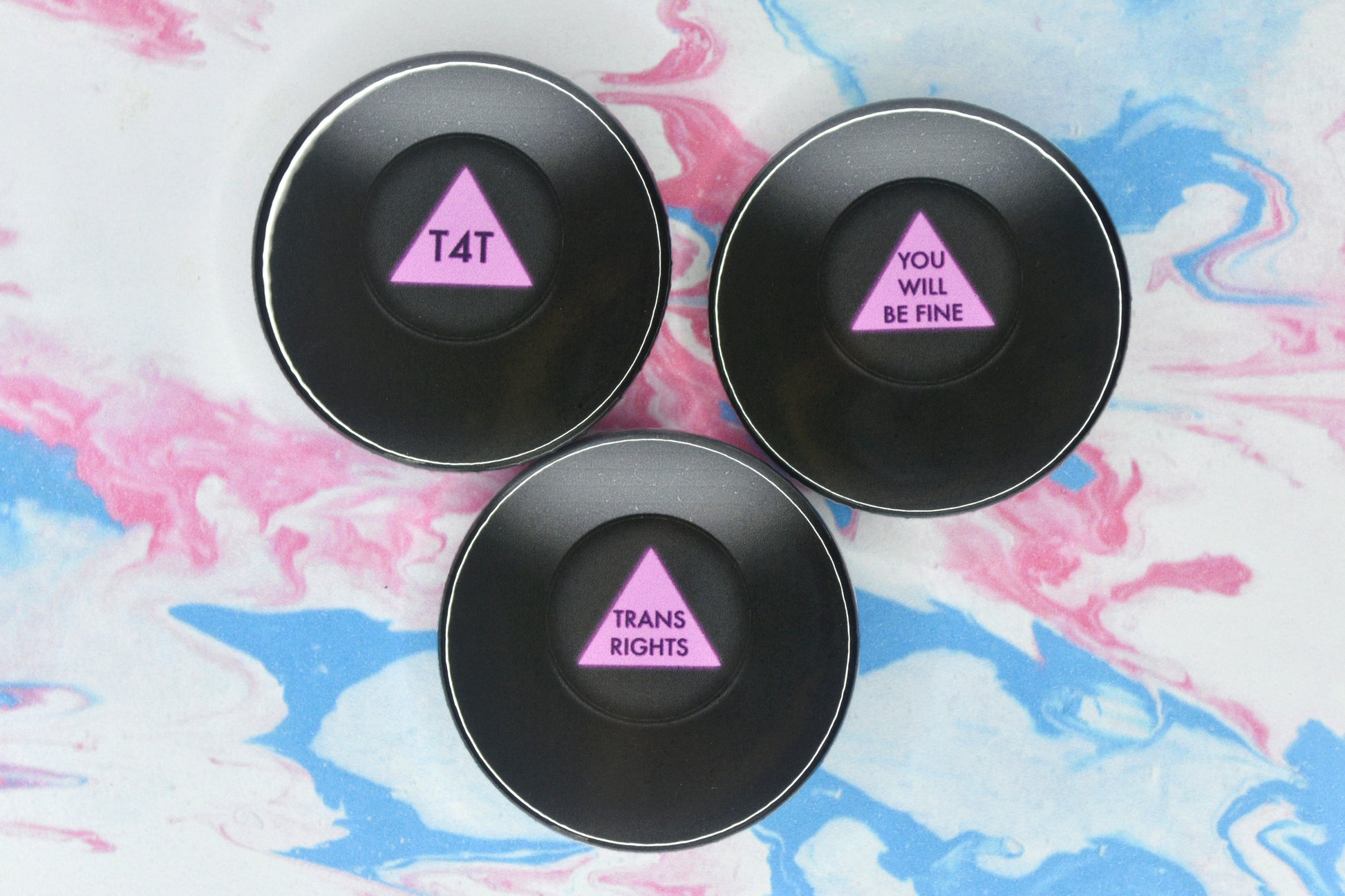 Three circular badges in size medium, each featuring an illustration of a black magic 8 ball which takes up the full badge. There is a small circular window in the centre which shows a pink triangle with the message of the ball written on it in darker pink text. The text on each reads "T4T", "You Will Be Fine" and "Trans Rights"