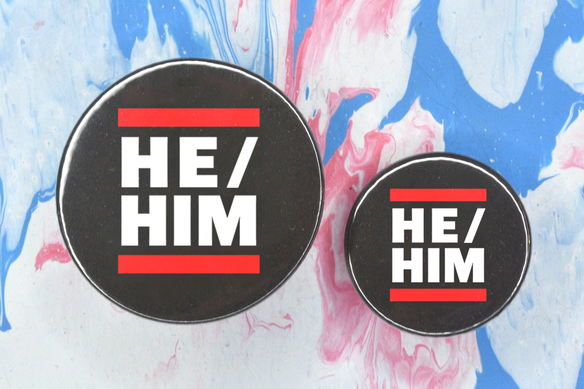 Two circular pin badges in sizes medium and small each with a black background, two rows of bold white upper case text and a solid red line above and below the text. The top row reads "he" with a forward slash and the bottom row reads "him".