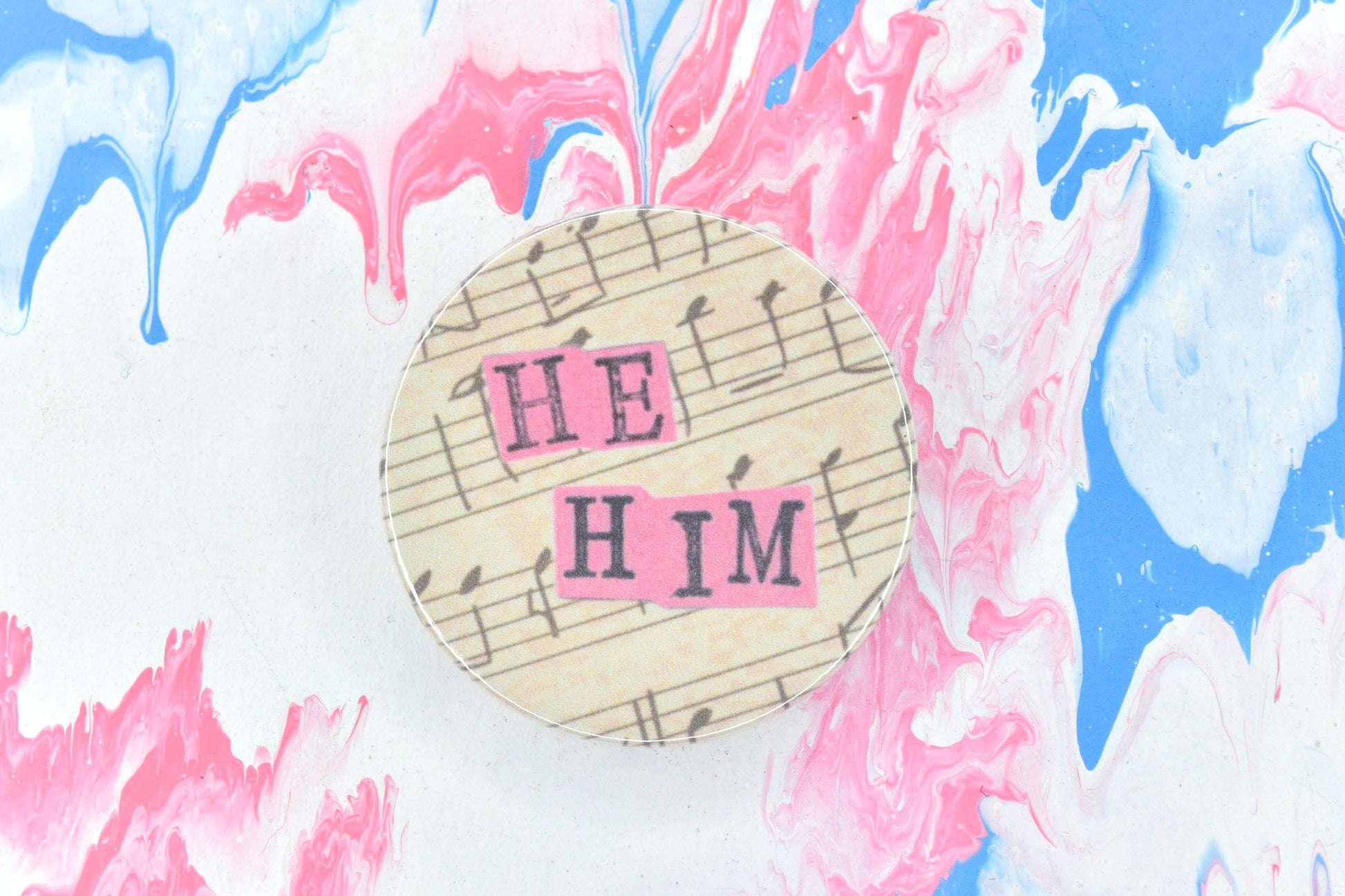 A pin badge featuring an off-white background with bars of music in black ink. Overlaid are hand cut pieces of pastel pink paper with hand stamped uppercase black lettering reading "he" and "him".