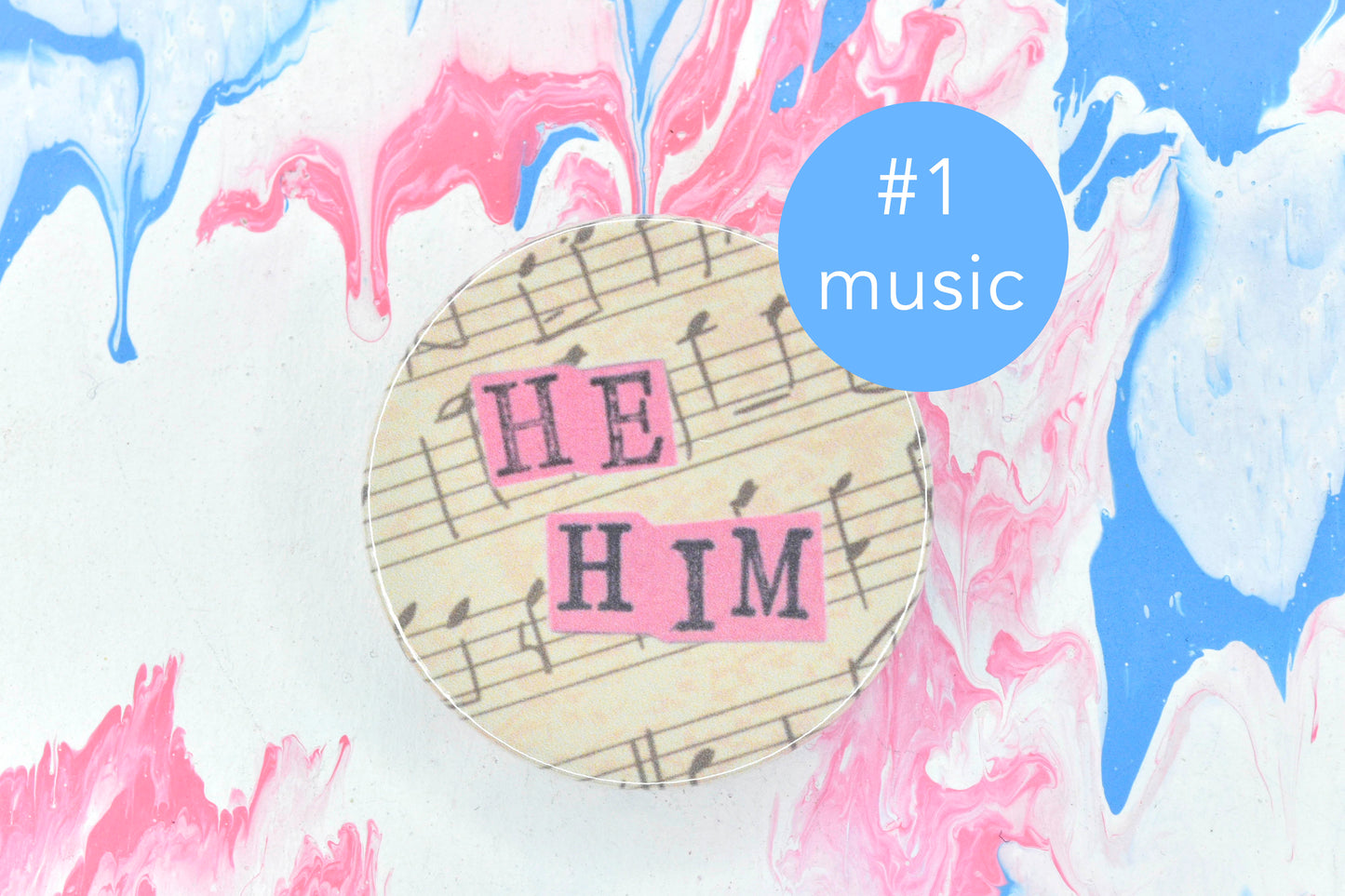 A pin badge featuring an off-white background with bars of music in black ink. Overlaid are hand cut pieces of pastel pink paper with hand stamped uppercase black lettering reading "he" and "him". There is a circular blue label next to the badge with white text reading "number 1, music".