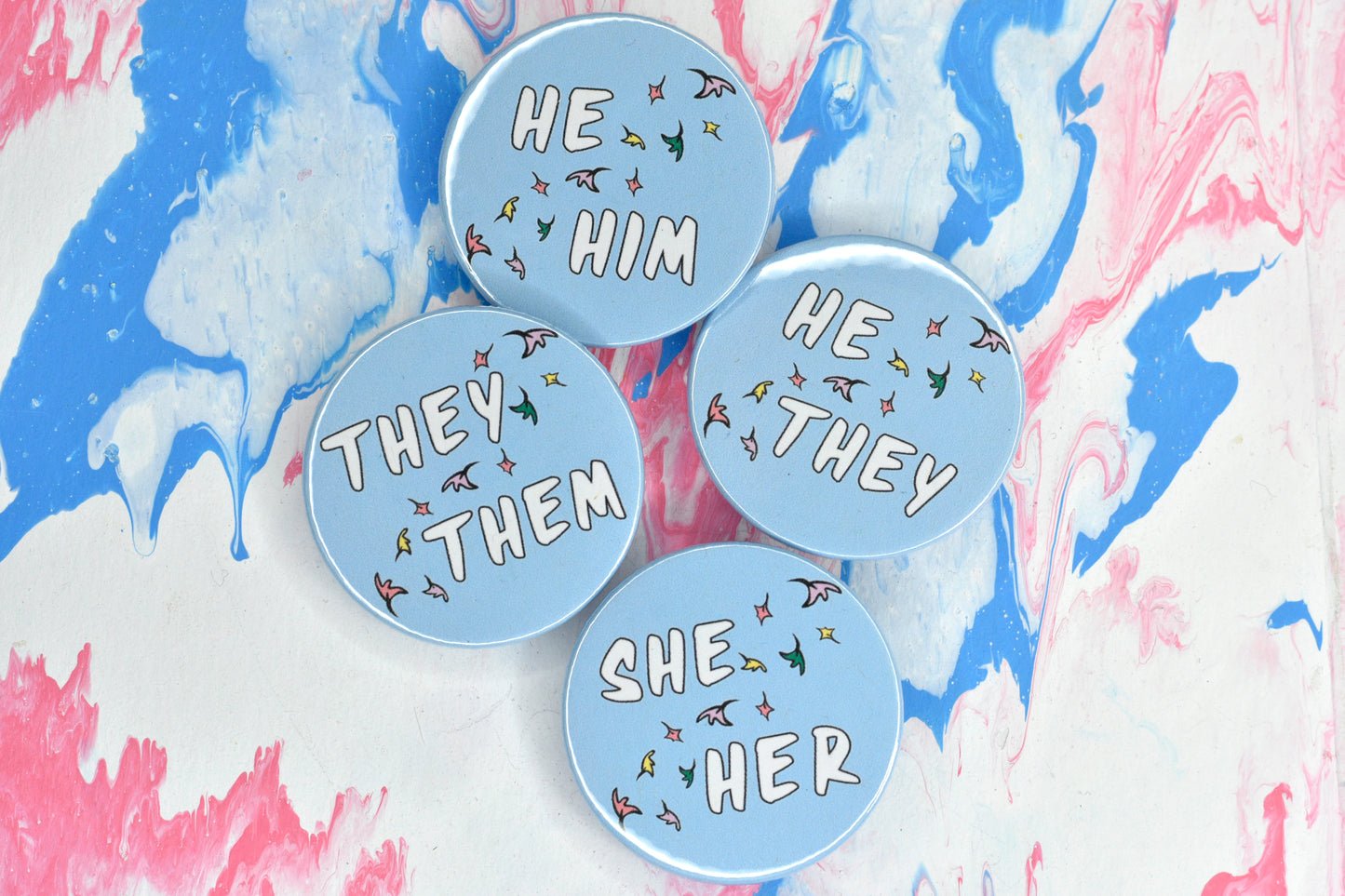 A photo of four round pin badges. Each has a pastel blue background and a set of pronouns in uppercase white text. The pronouns are separated by a diagonal band of pastel coloured comic book style leaves. The pronouns on the pins are "he him", "he/they" "she her" and "they them".