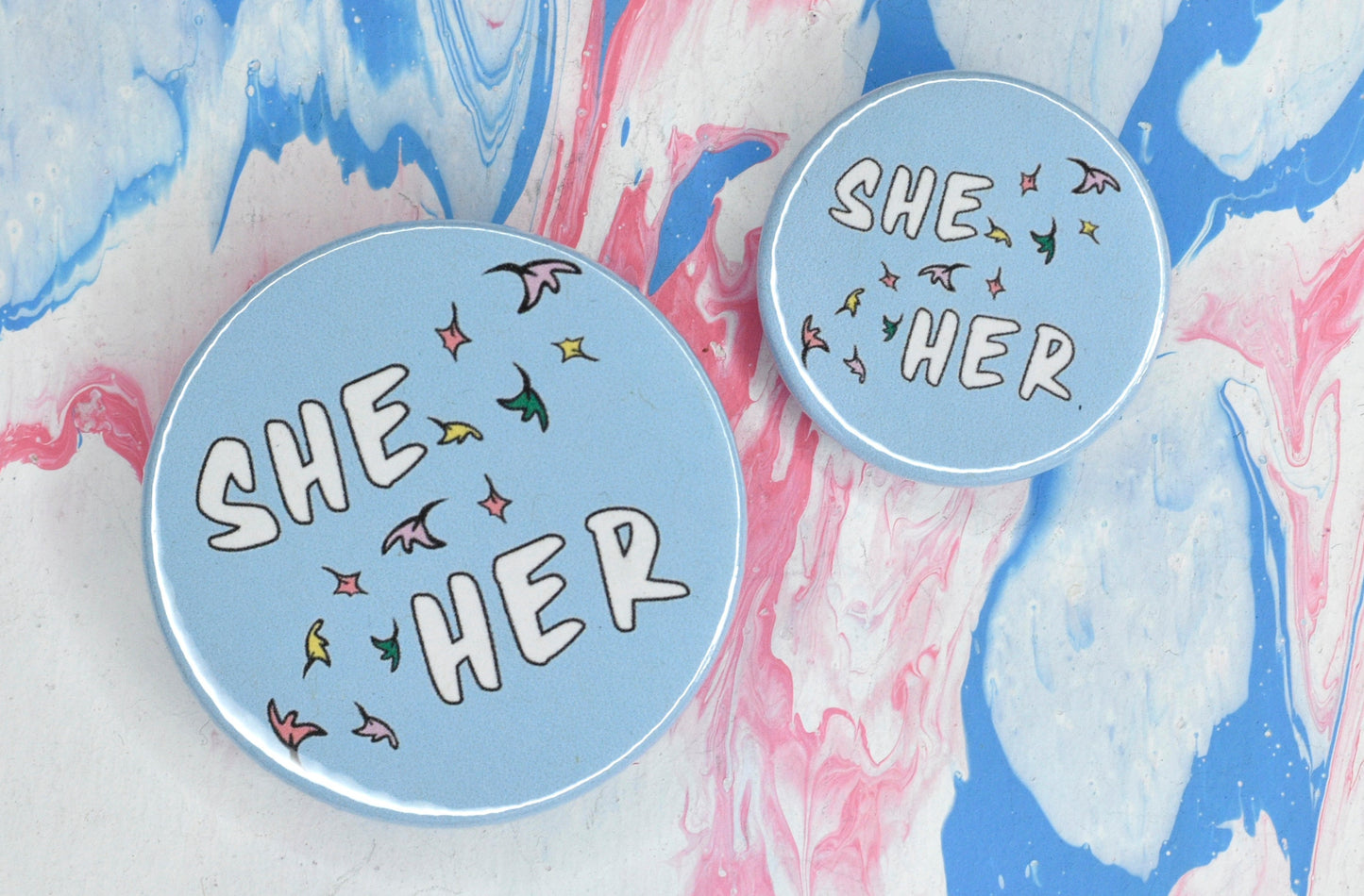 Two circular pronoun pin badges in sizes medium and small, each with a pastel blue background and a set of pronouns in uppercase white text. The pronouns are separated by a diagonal band of pastel coloured comic book style leaves. The pronouns shown are "she/her".